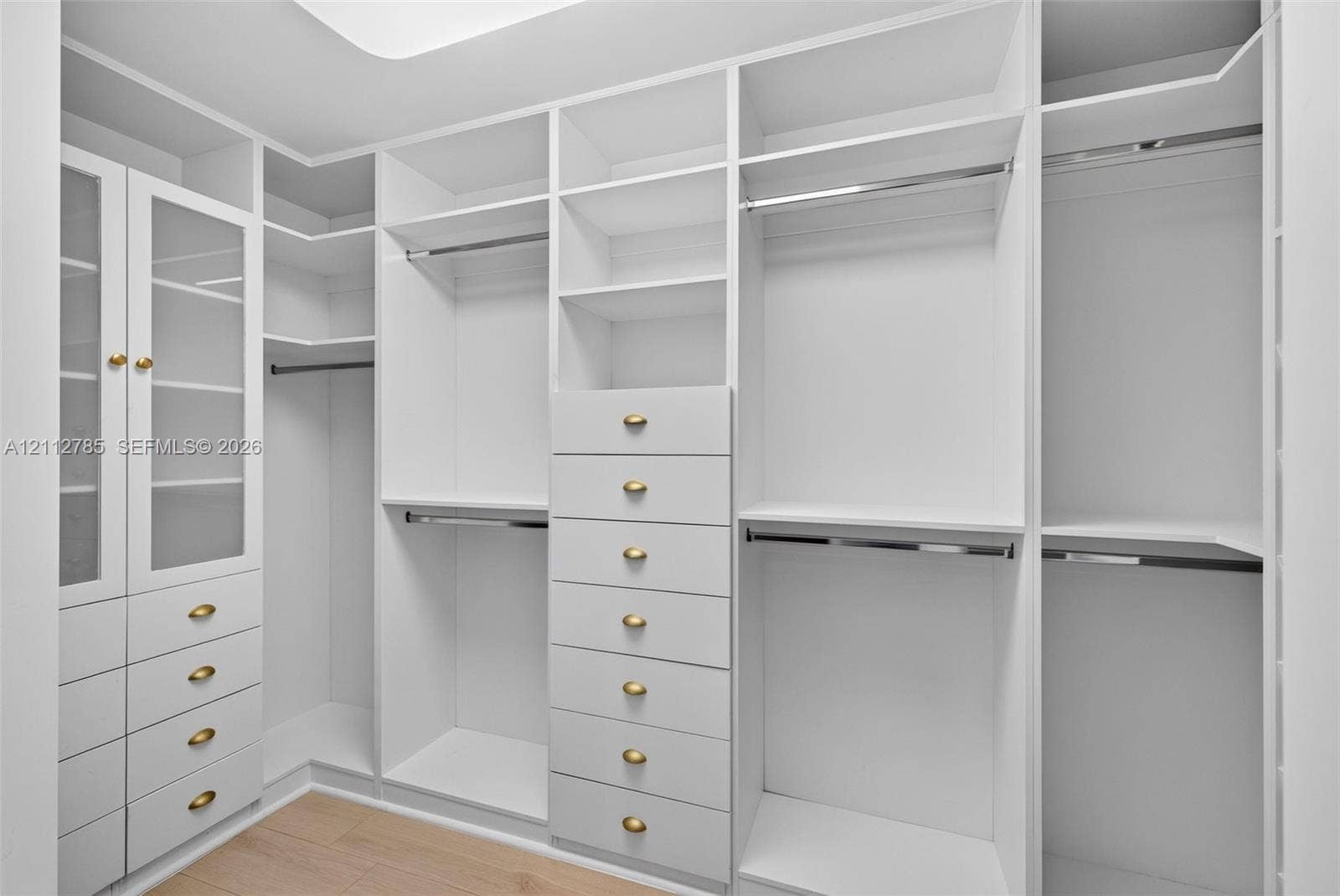 16425 Collins Avenue, #2016, Sunny Isles Beach, FL 33160 Custom walk-in closet featuring white built-in cabinetry, glass-front upper storage, multiple hanging rods, and integrated drawers with metallic hardware finished with wood-finish flooring