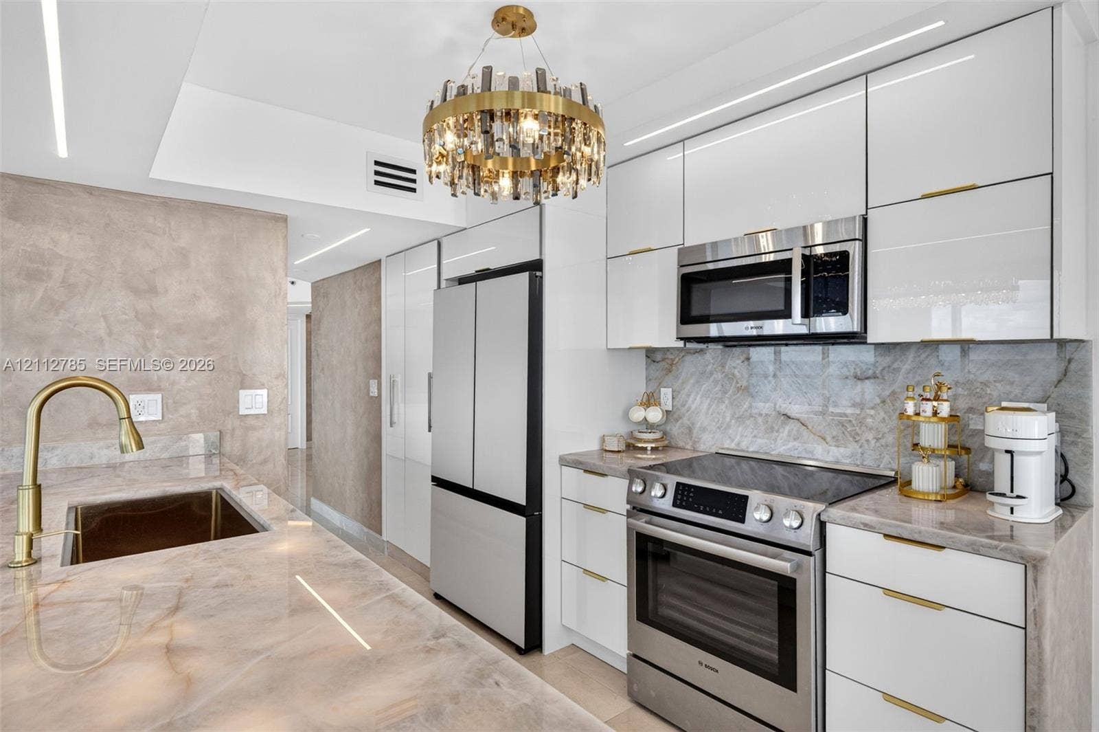 16425 Collins Avenue, #2016, Sunny Isles Beach, FL 33160 Glossy white cabinetry with gold-tone hardware, stone-finish countertops and backsplash, professional-grade stainless steel appliances, and a circular crystal chandelier