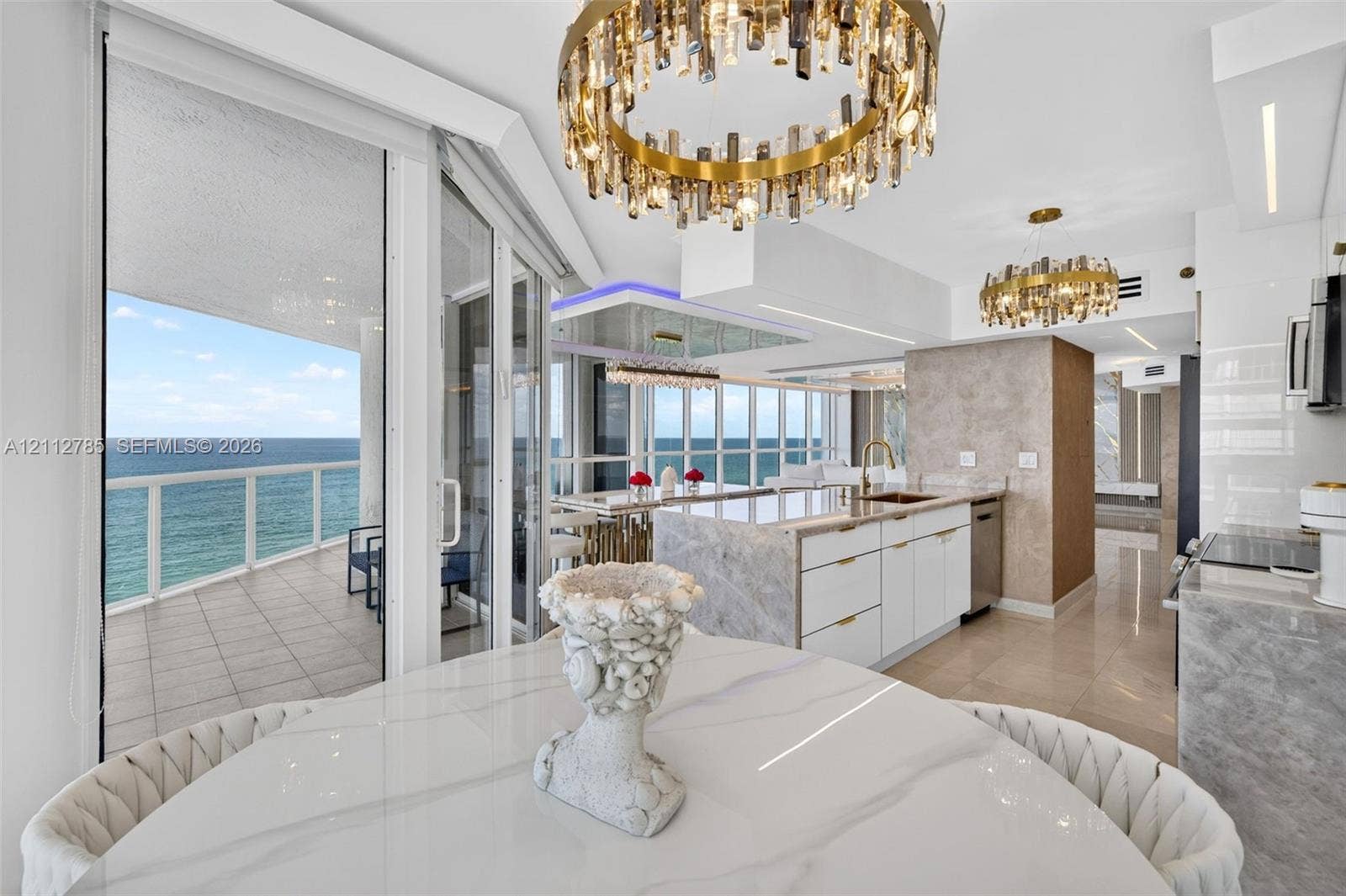 16425 Collins Avenue, #2016, Sunny Isles Beach, FL 33160 Open-concept kitchen featuring an island with stone-finish countertops, custom white cabinetry, gold-tone hardware, multi-tiered crystal statement chandeliers, and floor-to-ceiling glass doors opening to a tiled balcony