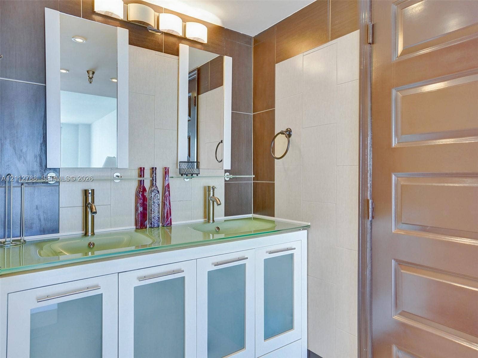 Double vanity with glass countertop, integrated sinks, white cabinetry with frosted glass inserts, metallic hardware, and dual vertical wall-mounted mirrors