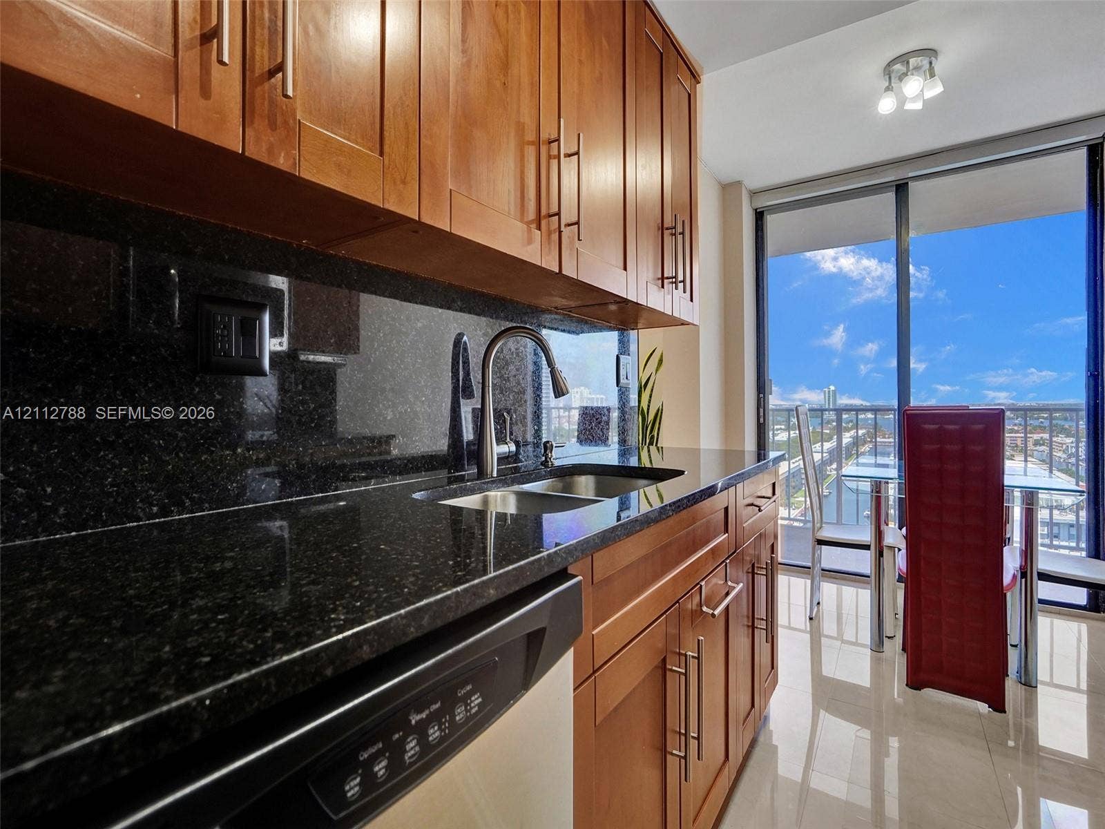 Kitchen featuring warm-tone wood cabinetry, dark stone countertops, stainless steel double-basin sink with gooseneck faucet, light-reflective tile flooring, and floor-to-ceiling glass doors