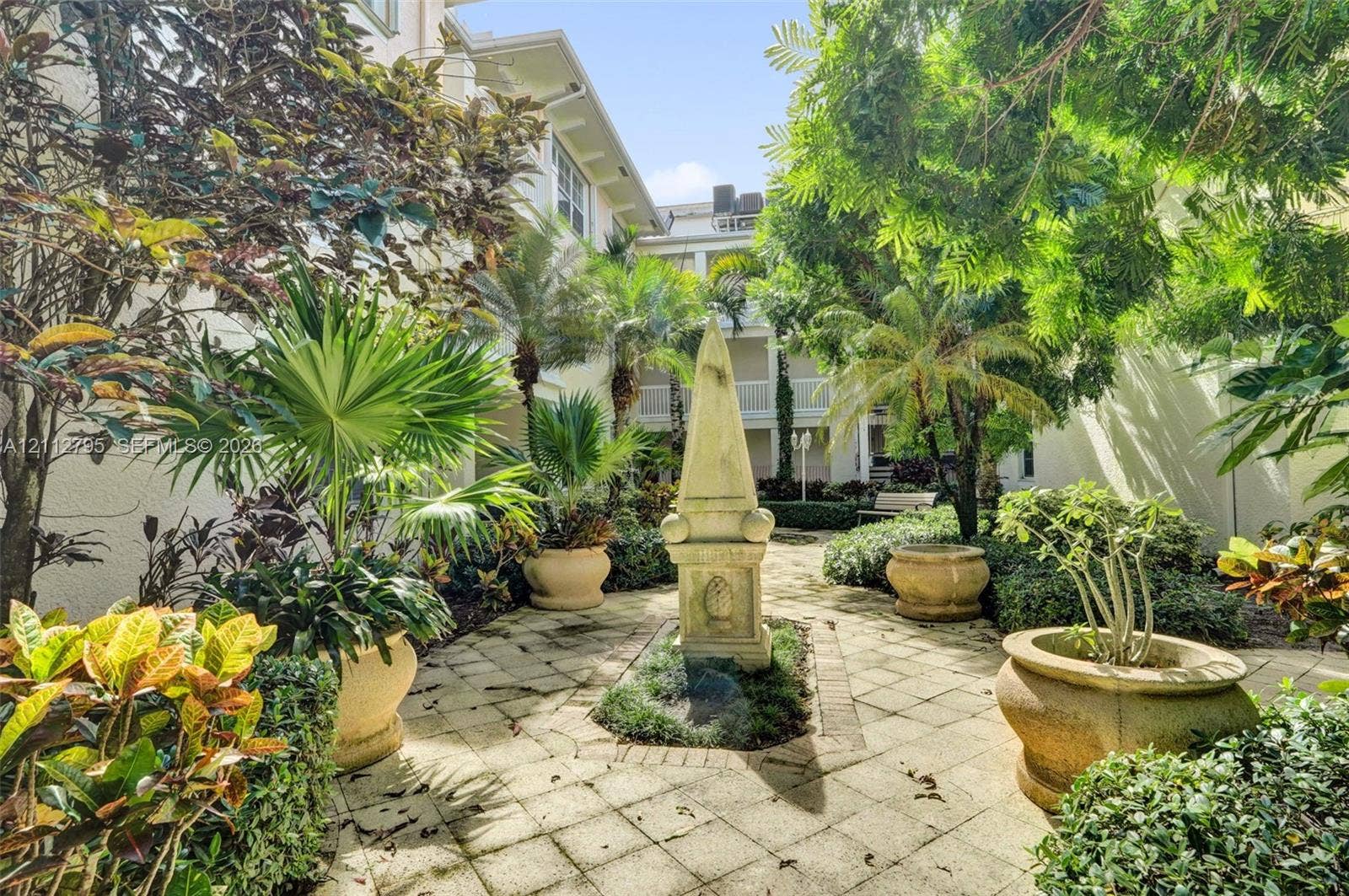 275 Murcia Drive, #205, Jupiter, FL 33458 Courtyard featuring stone paver flooring, central stone obelisk monument, mature trees, ornamental palms, and oversized stone planters