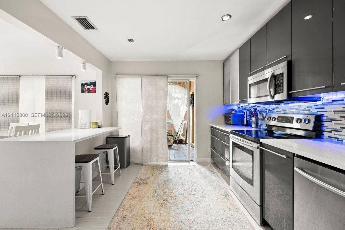 599 Racquet Club Road, #68-17, Weston, FL 33326 Kitchen featuring high-gloss charcoal cabinetry, stainless steel appliances, quartz countertops, a stone-tile backsplash, and a breakfast bar