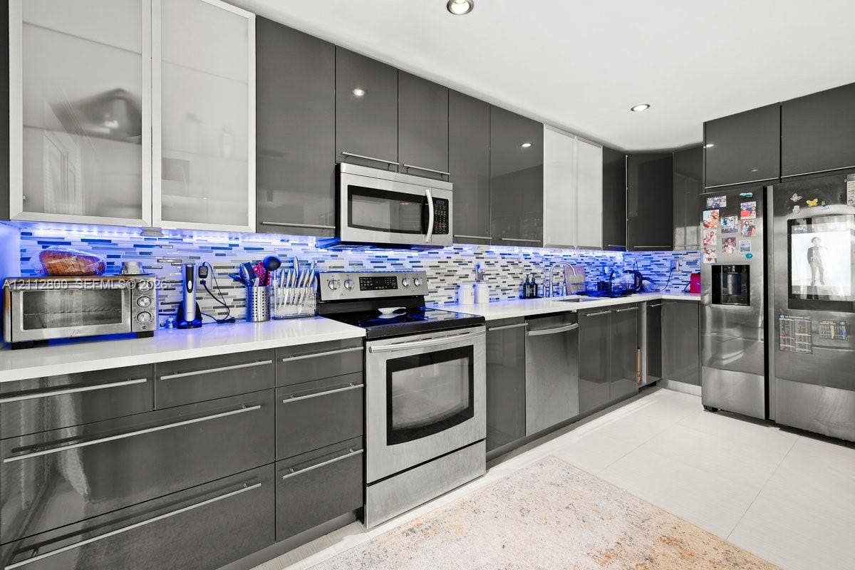 599 Racquet Club Road, #68-17, Weston, FL 33326 Kitchen featuring high-gloss charcoal cabinetry, glass-front upper cabinets, stainless steel appliances, a multi-tone glass tile backsplash, and quartz countertops