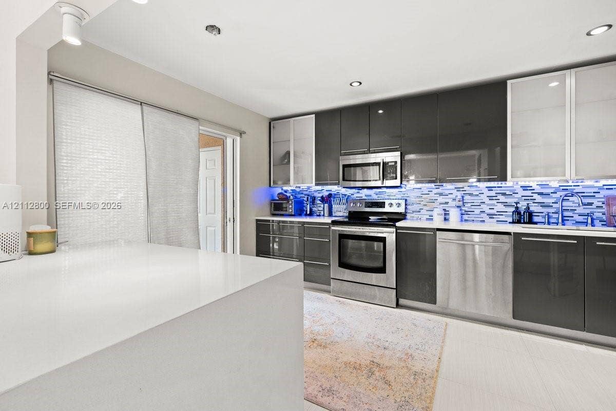 599 Racquet Club Road, #68-17, Weston, FL 33326 Modern kitchen featuring high-gloss charcoal cabinetry, stainless steel appliances, a white quartz waterfall-edge island, glass-front upper cabinets, and a multi-toned mosaic tile backsplash