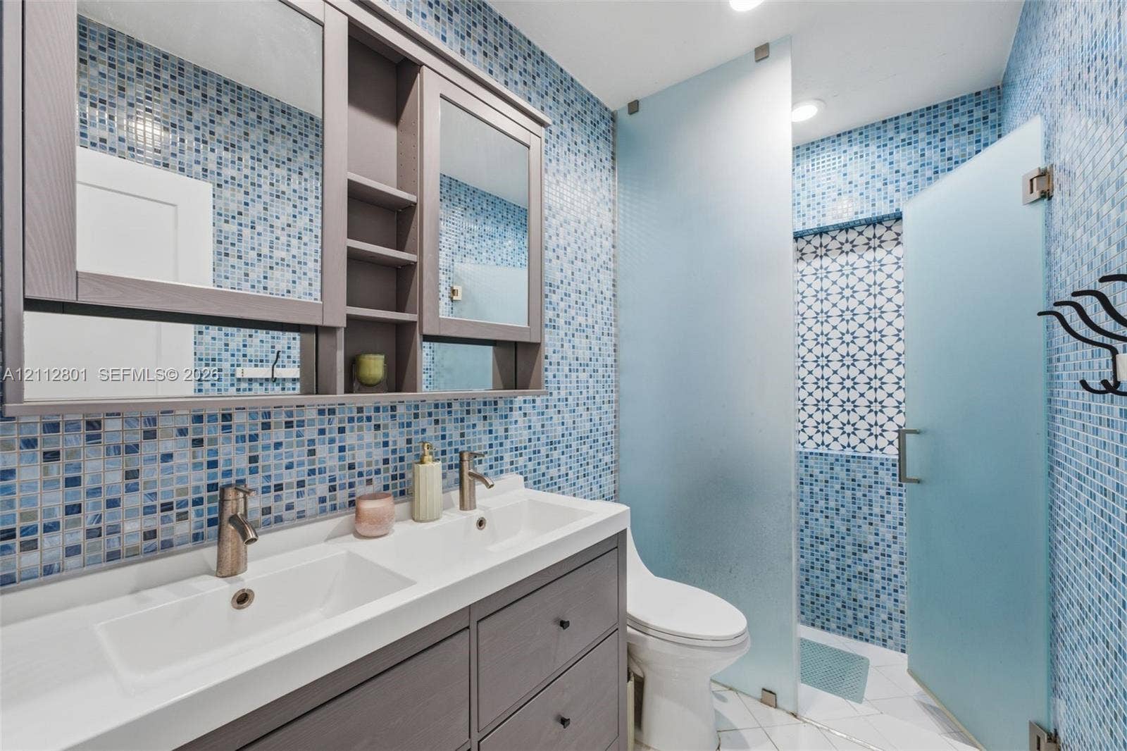 3241 SW 84th Court, Miami, FL 33155 Double vanity featuring a white integrated countertop with dual basins and matte-finish hardware, built-in medicine cabinets, full-height blue glass mosaic tile walls, and a walk-in shower enclosure with frosted glass panels