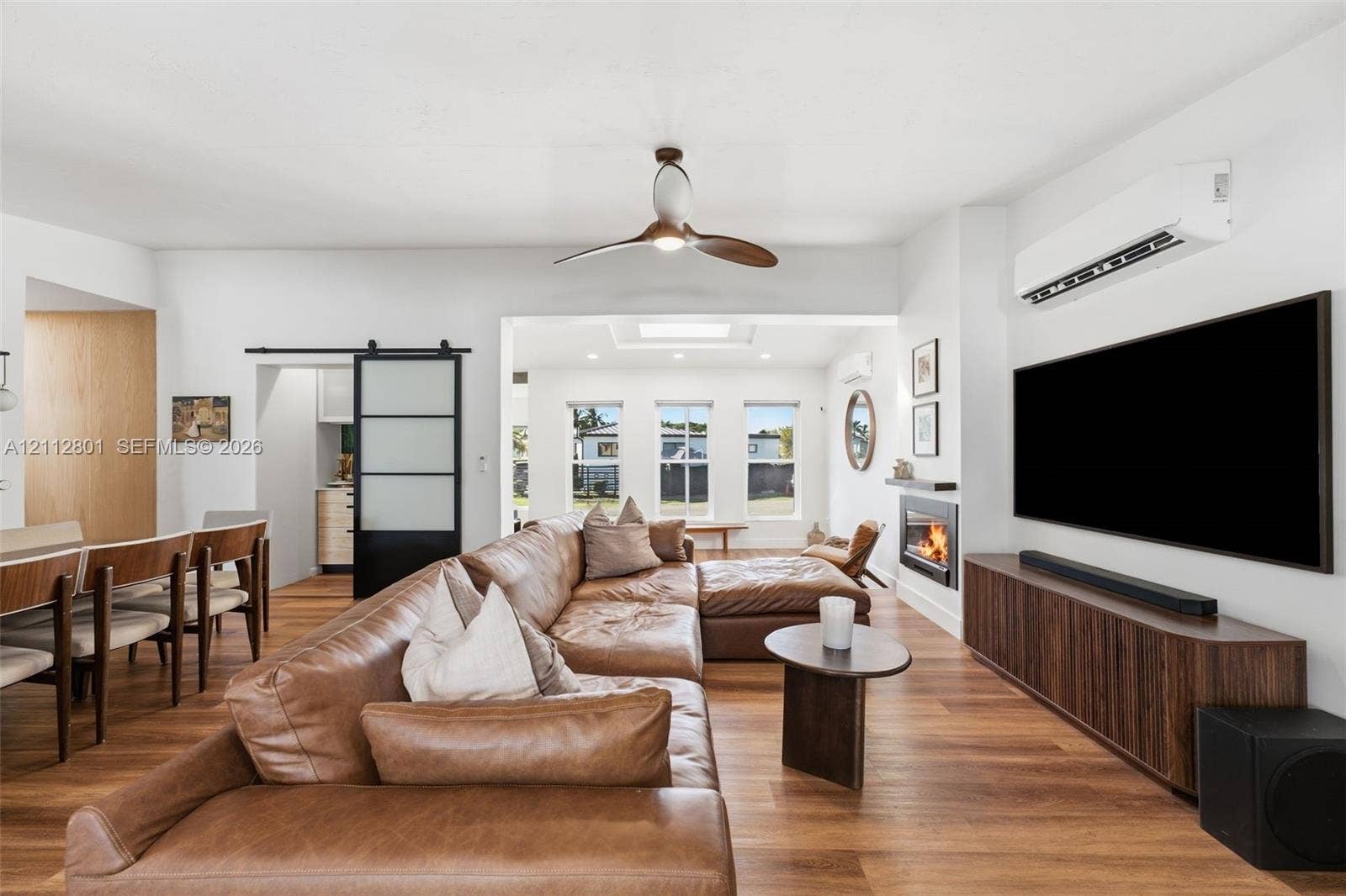 3241 SW 84th Court, Miami, FL 33155 Open-concept interior featuring wood-finish flooring, a contemporary ceiling fan, a wall-mounted fireplace, a black-framed sliding barn door with frosted panels, and a wall-mounted mini-split HVAC unit