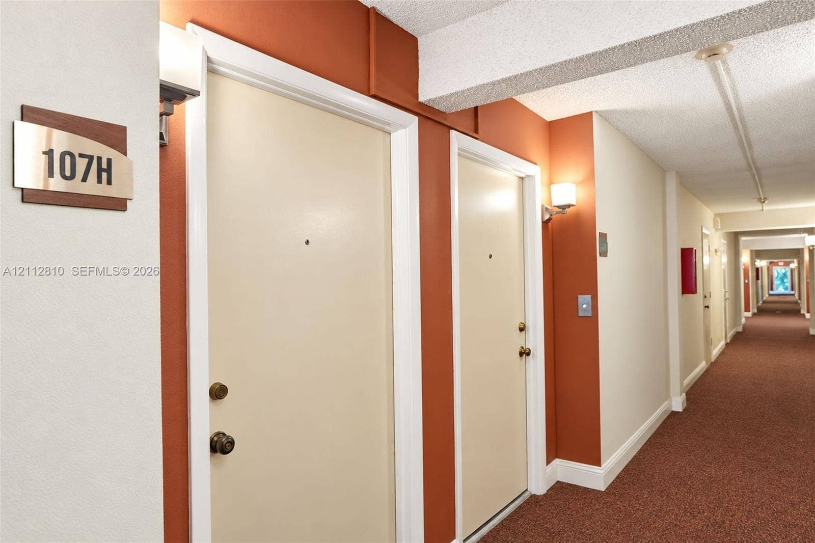 1920 NE 1st Terrace, #107H, Wilton Manors, FL 33305 Interior hallway featuring wall-mounted light fixtures, carpeted flooring, and neutral-toned doors with metallic hardware