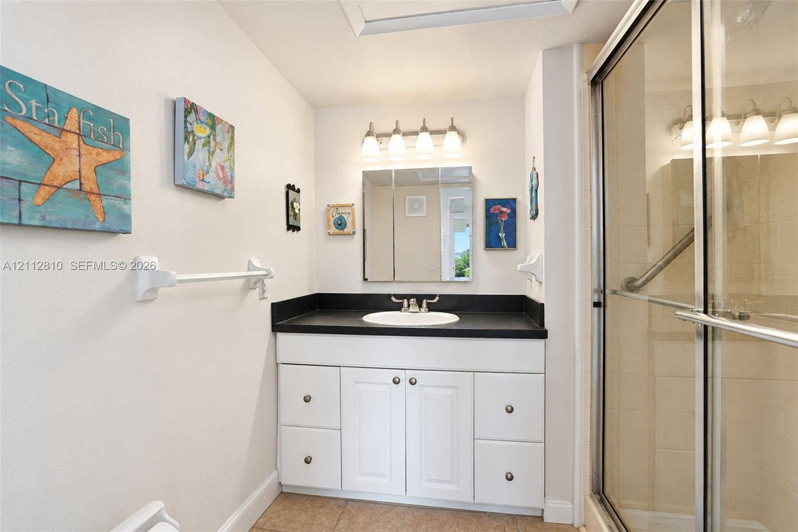 1920 NE 1st Terrace, #107H, Wilton Manors, FL 33305 Bathroom vanity featuring white cabinetry, a dark countertop, an integrated sink, a triple-light fixture, and a tri-fold medicine cabinet, paired with a glass-enclosed shower