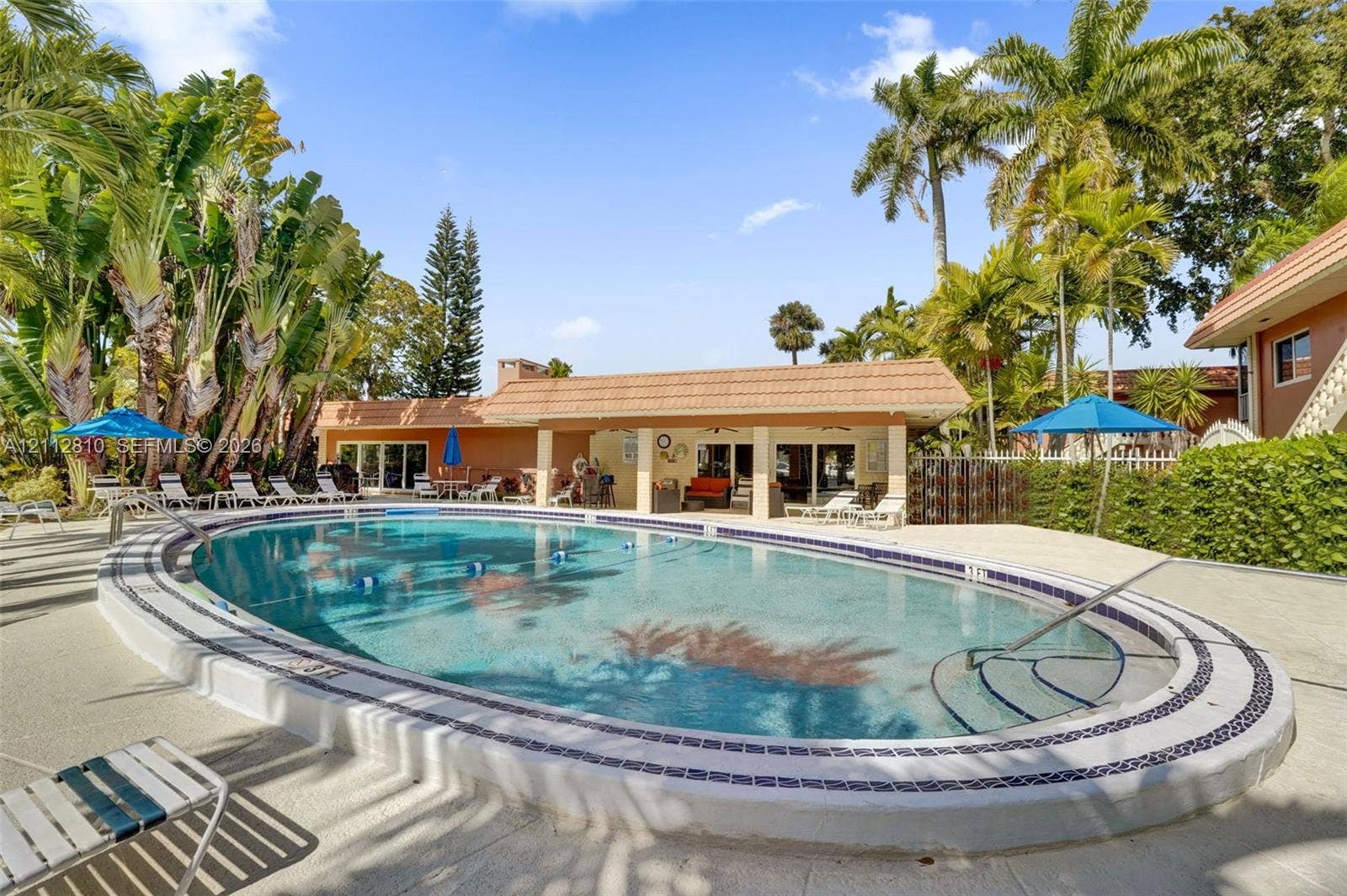 1920 NE 1st Terrace, #107H, Wilton Manors, FL 33305 Oval swimming pool with tile border, recessed steps, and light-colored concrete decking, situated within a landscape featuring mature palm trees and a covered patio structure with tile roofing