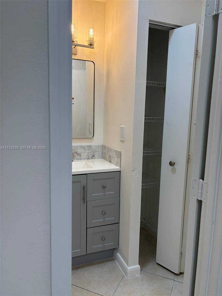 508 NW 43rd Place, Deerfield Beach, FL 33064 Gray vanity cabinet with a white countertop, tiled backsplash, rectangular mirror with metallic frame, two-light fixture, and built-in closet with wire shelving