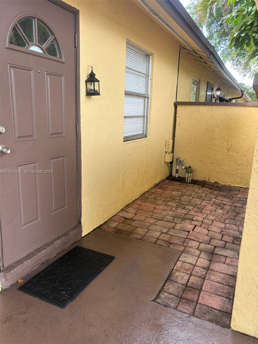 508 NW 43rd Place, Deerfield Beach, FL 33064 Exterior entryway featuring a solid-paneled door with an arched glass insert, exterior lantern-style wall lighting, and brick paver flooring