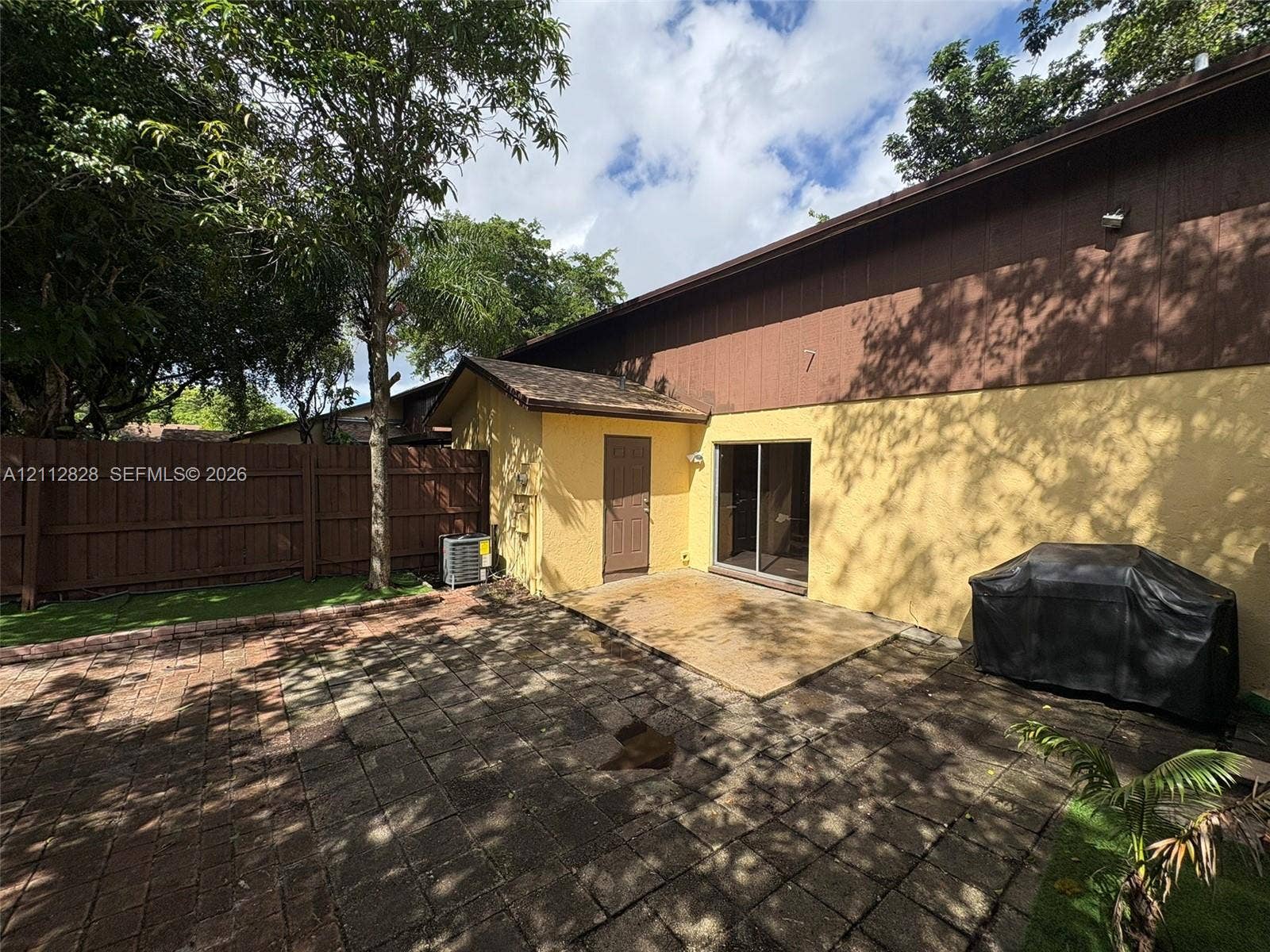 508 NW 43rd Place, Deerfield Beach, FL 33064 Rear exterior featuring a paved stone patio, dual-tone stucco and wood siding, a sliding glass door, and a privacy-fenced yard