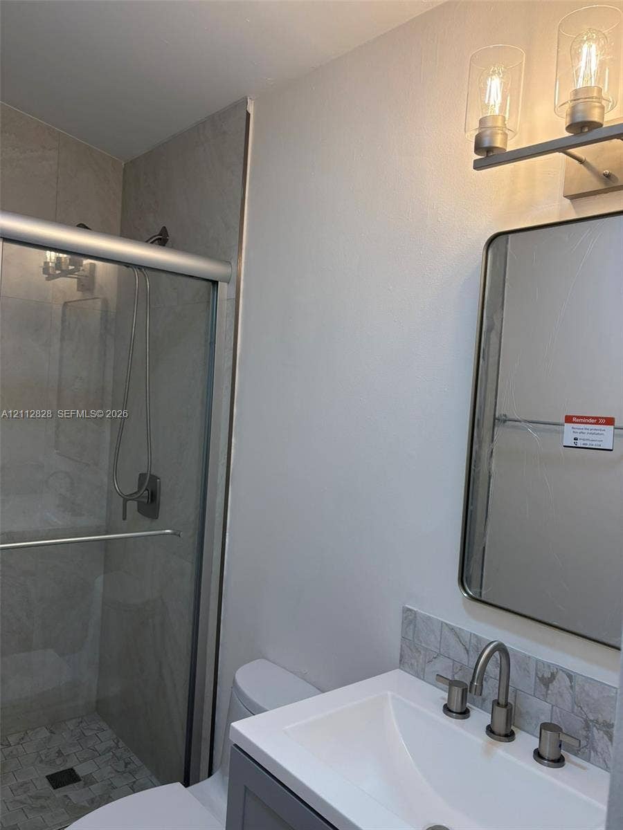508 NW 43rd Place, Deerfield Beach, FL 33064 Shower with sliding glass doors and tiled surround, vanity with stone-style backsplash, integrated sink, metal wall-mounted light fixture, and framed mirror