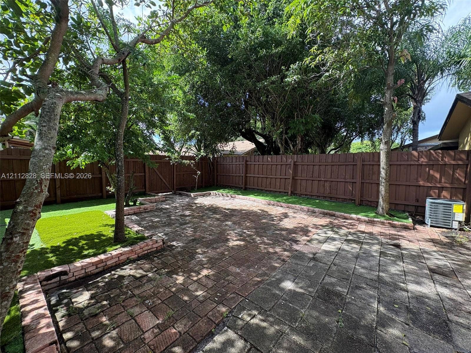 508 NW 43rd Place, Deerfield Beach, FL 33064 Rear yard featuring a paver patio, brick retaining walls, synthetic turf, wood privacy fencing, and mature trees