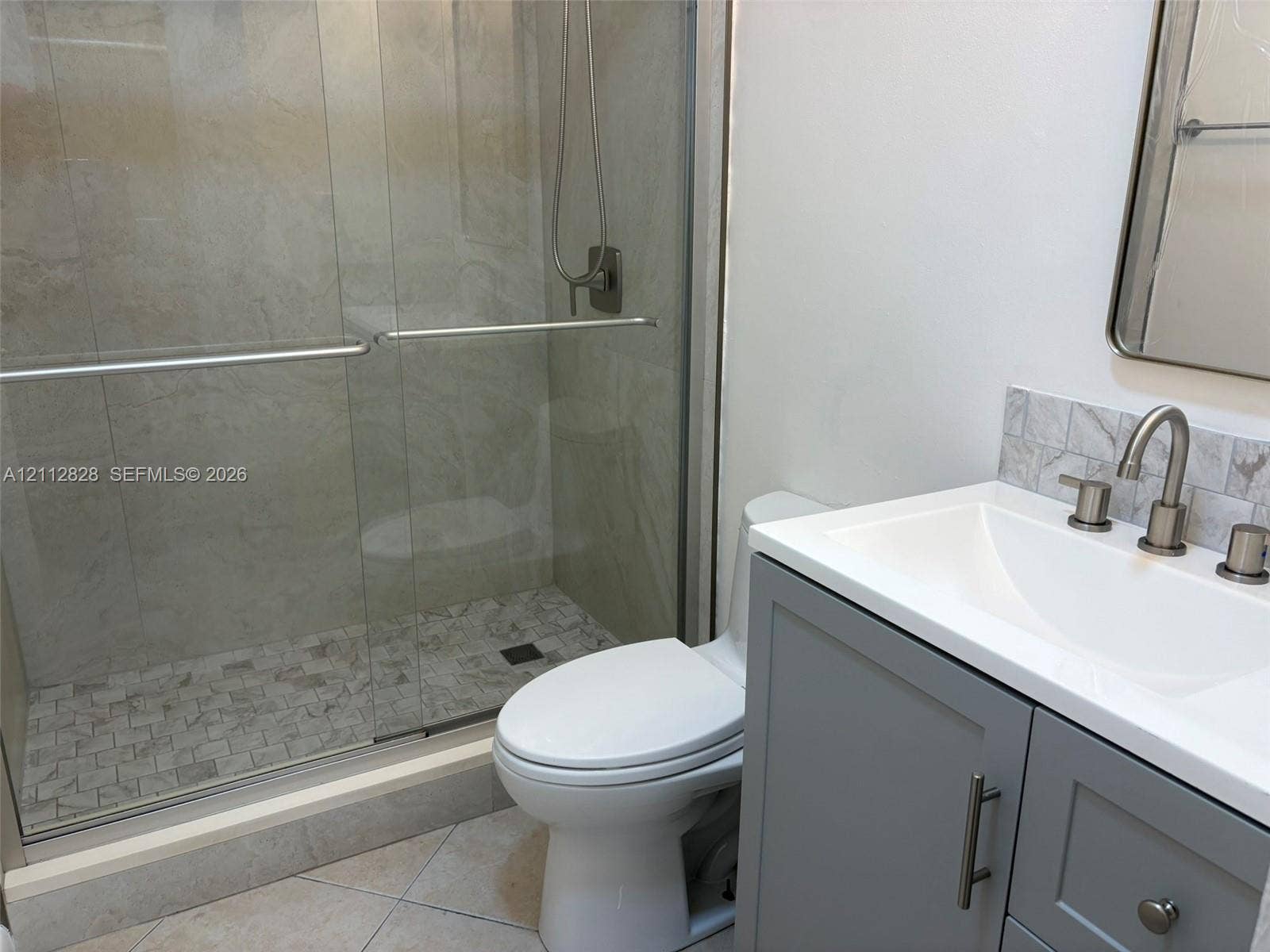 508 NW 43rd Place, Deerfield Beach, FL 33064 Bathroom featuring a walk-in shower with sliding glass doors, a vanity with a white countertop and undermount sink, stone-finish backsplash, and a wall-mounted mirror