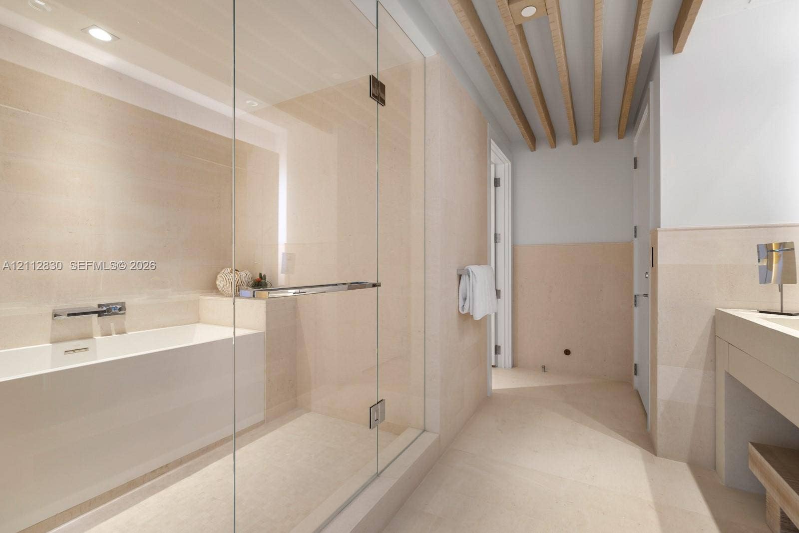 Bathroom featuring a glass-enclosed shower, soaking tub with wall-mounted faucet, neutral stone-finish wall and floor tile, and exposed wooden ceiling beams