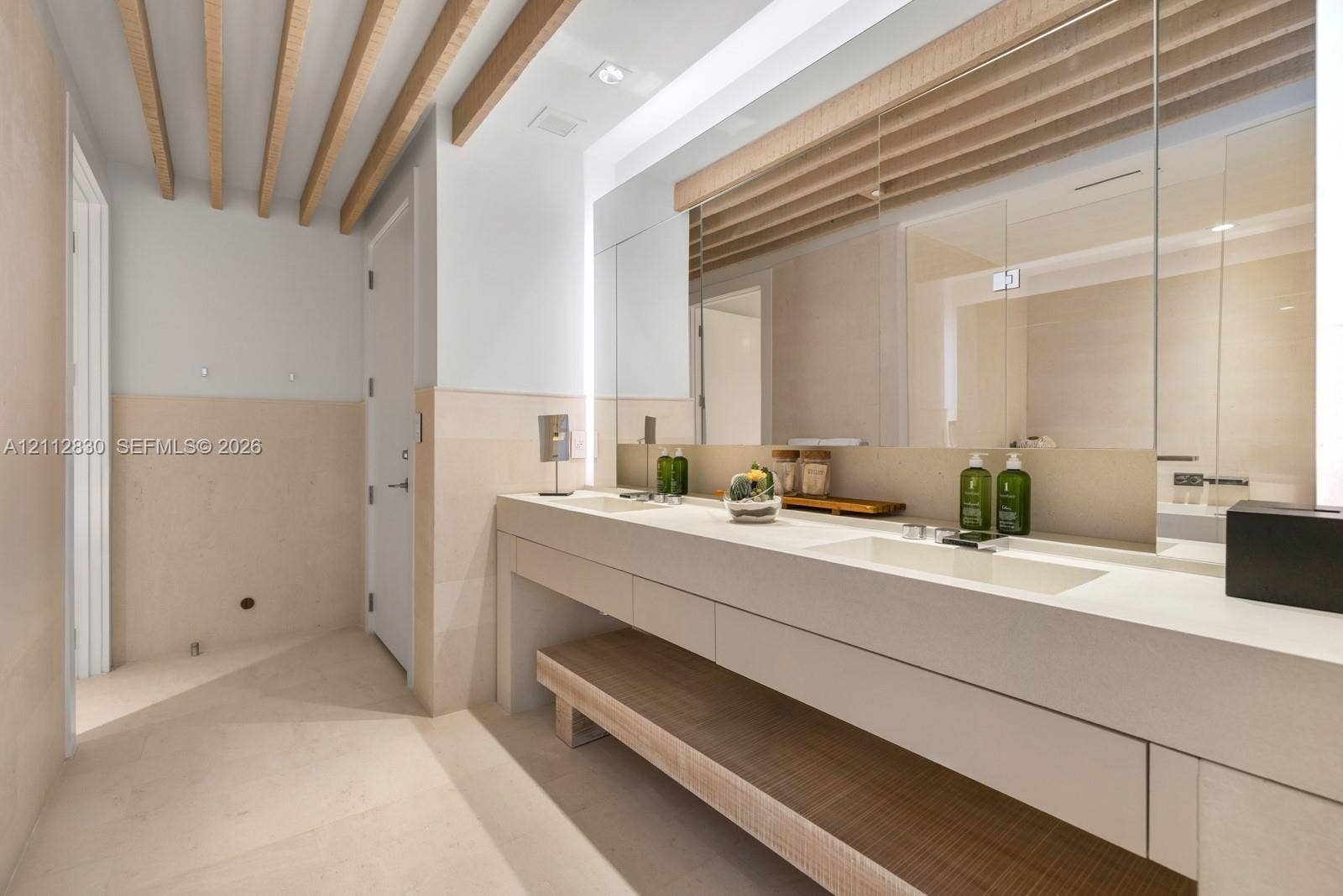Dual vanity with integrated sinks, stone-surface countertop, full-width mirror with integrated lighting, exposed wood ceiling beams, and stone-finish wall cladding