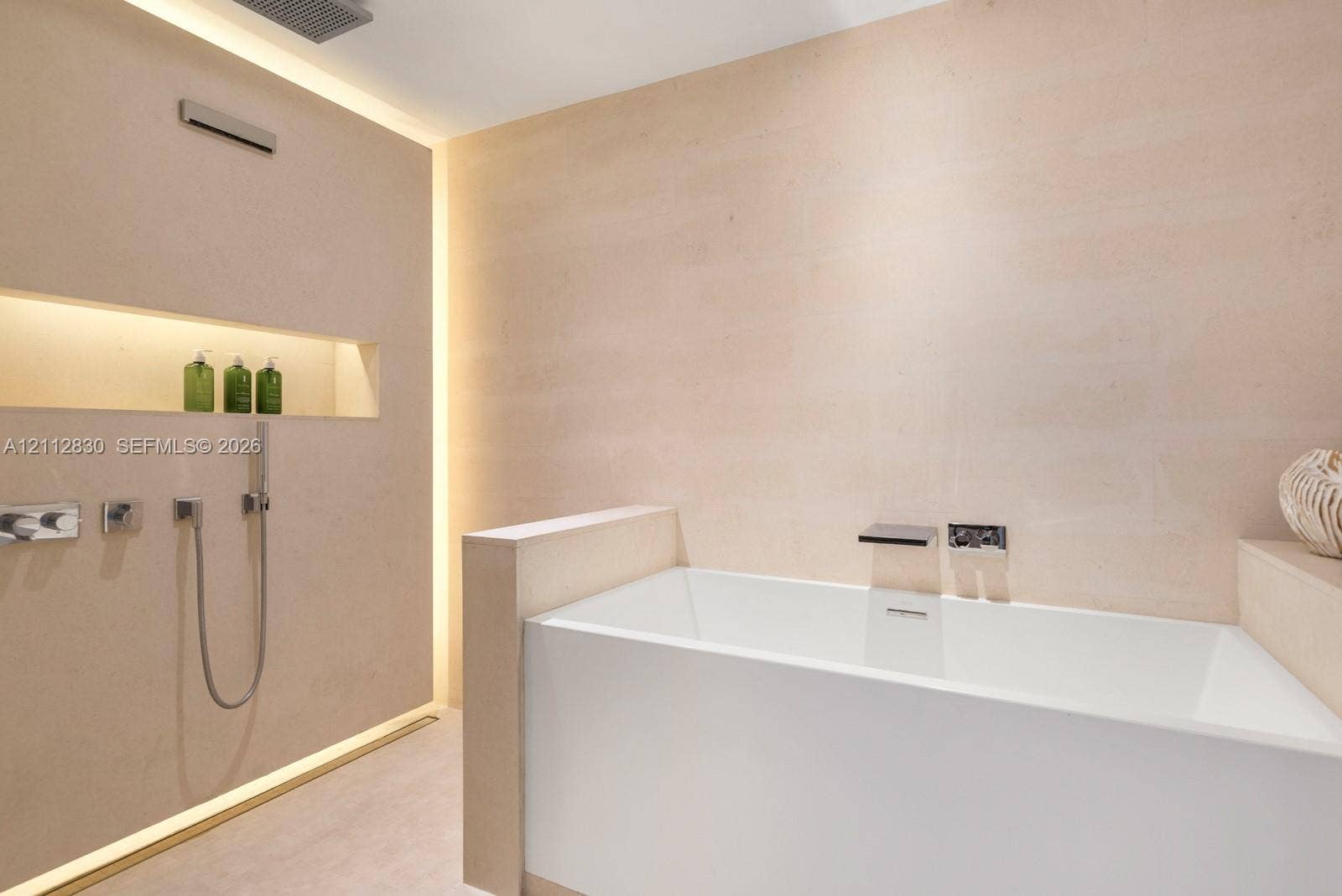 Freestanding deep-soaking bathtub with wall-mounted waterfall filler, integrated stone-finish wall cladding, recessed wall niche, concealed perimeter accent lighting, and polished chrome hand-held shower fixture