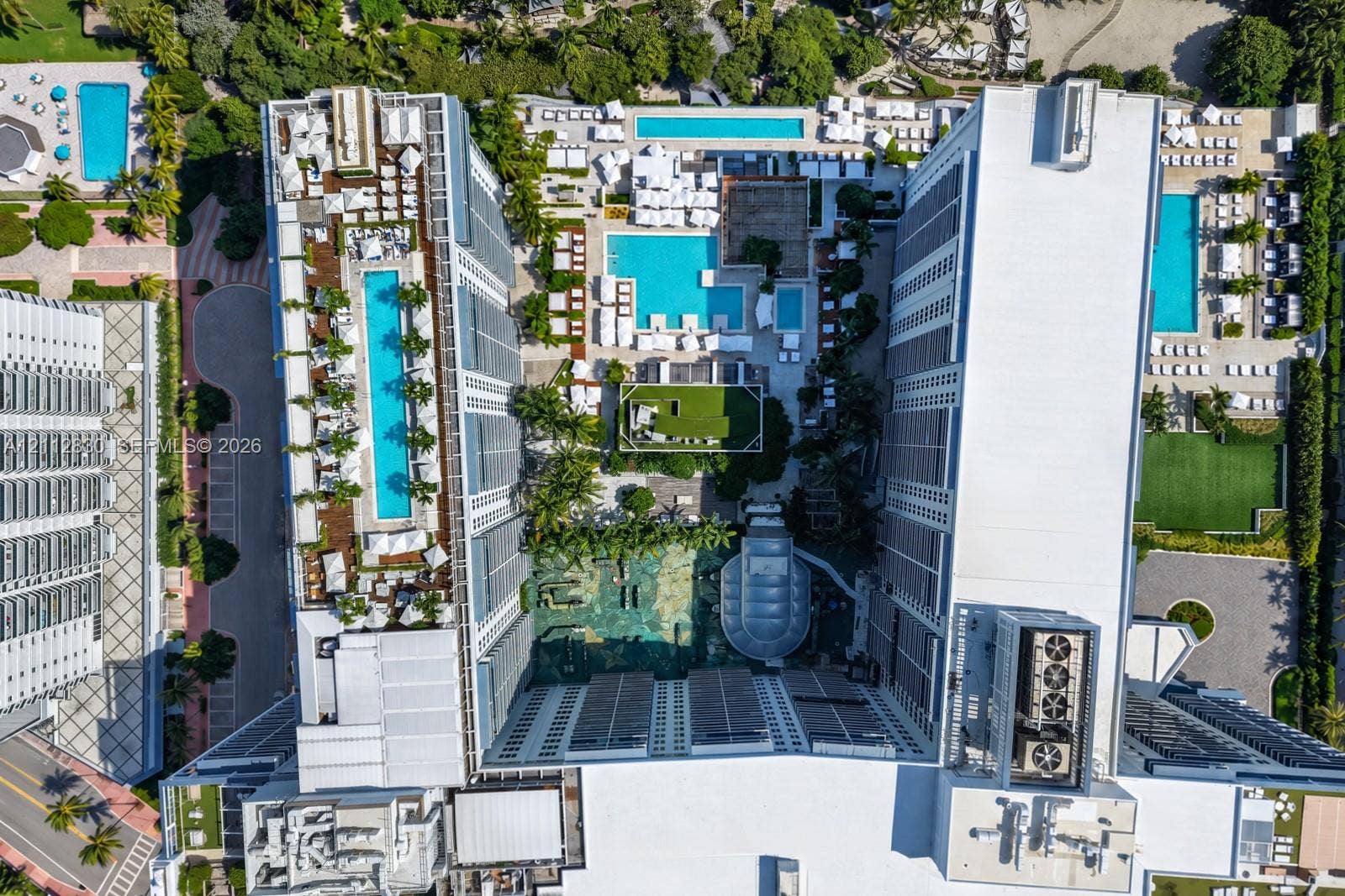 Aerial perspective of a multi-structure complex featuring multiple rectangular swimming pools, hardscaped recreation areas, integrated green space, and mature perimeter landscaping
