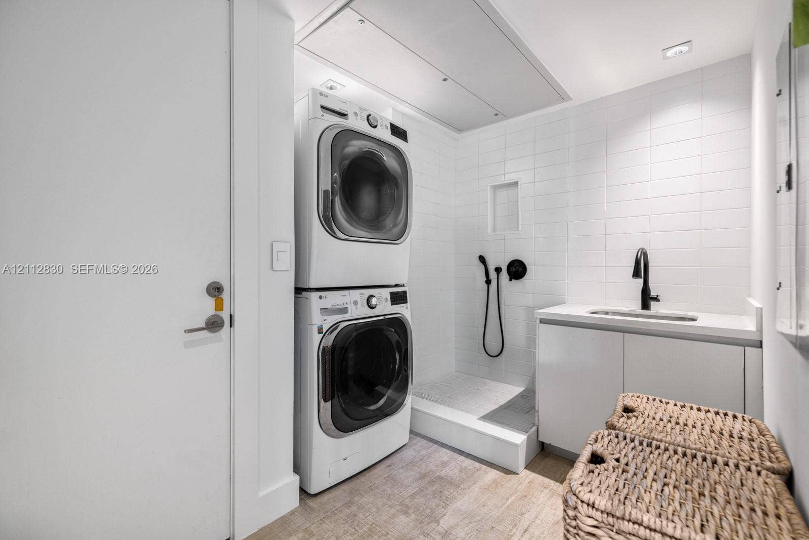 Laundry area featuring stacked appliances, tiled shower enclosure with handheld sprayer, built-in cabinetry, solid-surface countertop, and matte black faucet