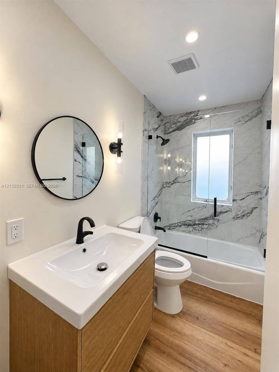 Modern vanity featuring a light wood-finish cabinet and white countertop, contrasting matte black plumbing fixtures, circular wall mirror, wood-finish flooring, and a shower-tub combination with full-height stone-patterned wall surrounds