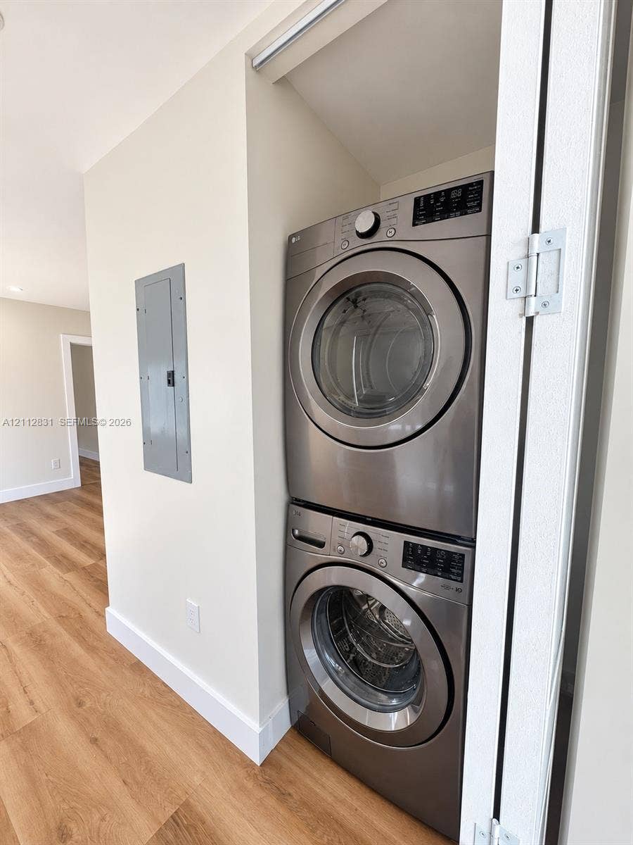 Stacked stainless steel laundry appliances situated within a dedicated closet, featuring wood-finish flooring and a wall-mounted electrical panel