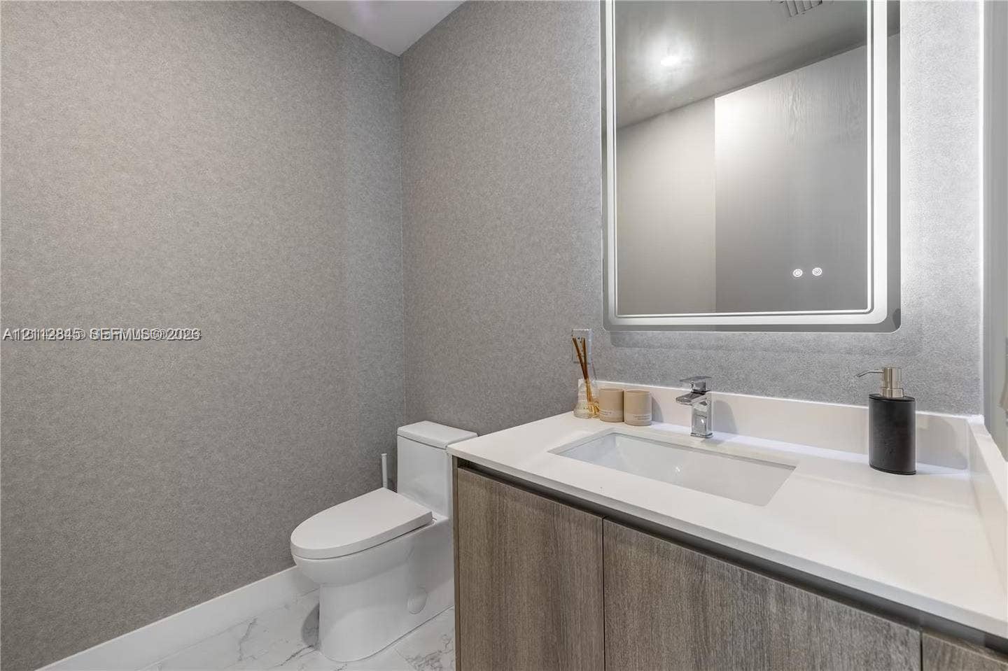 17550 Collins Avenue, #1202, Sunny Isles Beach, Sunny Isles Beach, FL 33160 Modern vanity with integrated rectangular sink and quartz countertop, complemented by a lighted rectangular mirror, textured wall covering, and stone-finish tile flooring