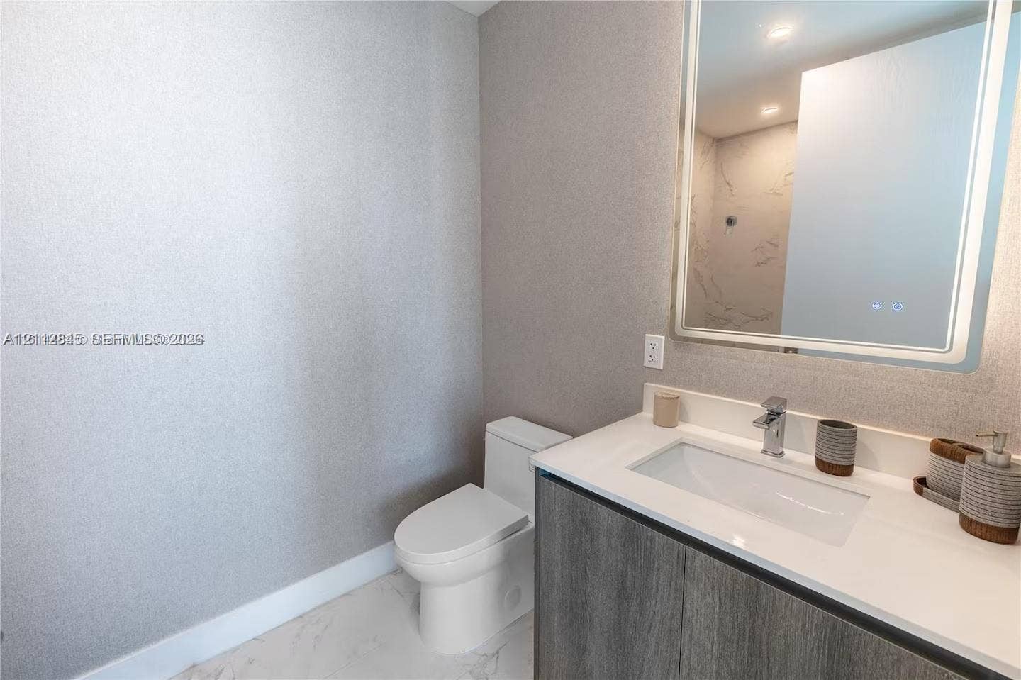 17550 Collins Avenue, #1202, Sunny Isles Beach, Sunny Isles Beach, FL 33160 Modern vanity featuring a charcoal-finish cabinet, rectangular basin, and stone-surface countertop, complemented by a large integrated backlit mirror and white tile flooring