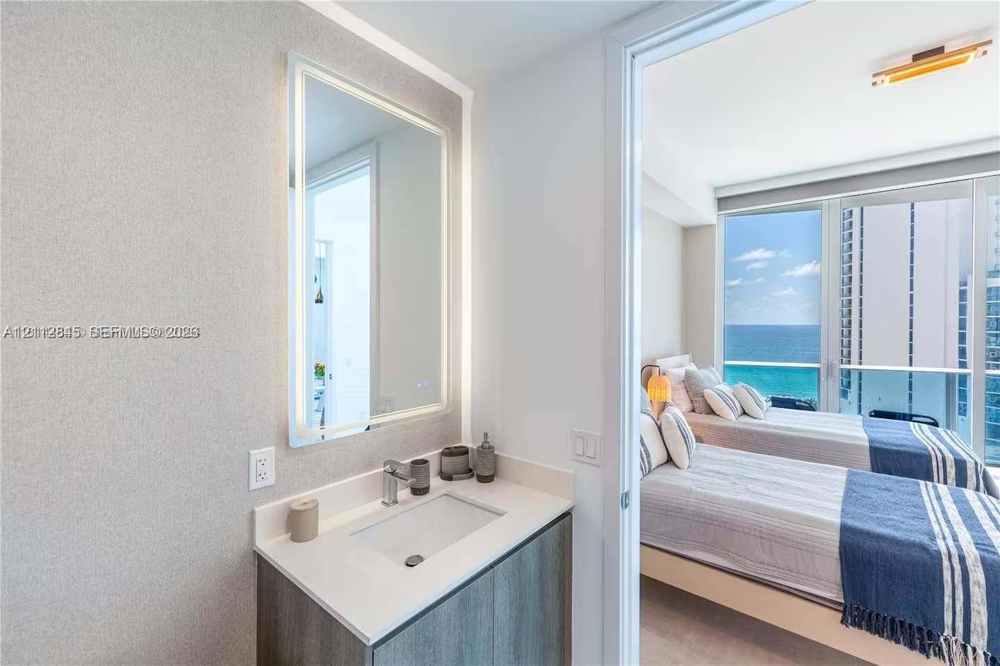 17550 Collins Avenue, #1202, Sunny Isles Beach, Sunny Isles Beach, FL 33160 Vanity featuring a stone-composite countertop, undermount sink, integrated LED-backlit mirror, and wood-finish cabinetry, paired with floor-to-ceiling glass windows and an exterior balcony