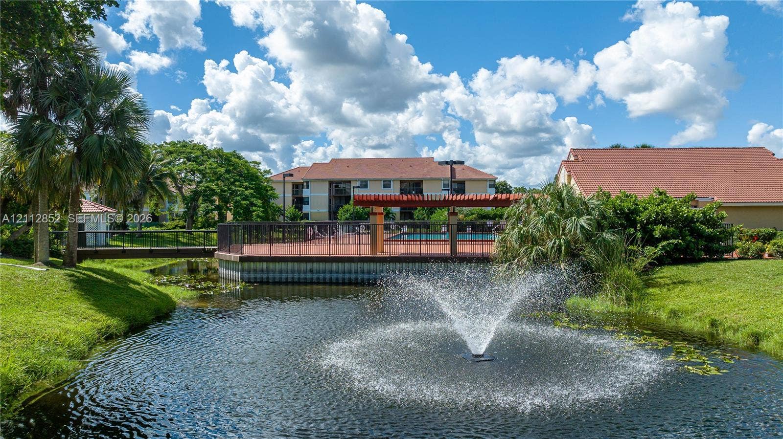 5741 Riverside Drive, #205A5, Coral Springs, Coral Springs, FL 33067 Community pond featuring a central fountain, manicured landscaping, pedestrian bridge, and paved pool deck with a wooden pergola