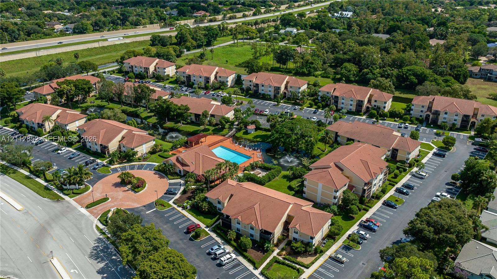 5741 Riverside Drive, #205A5, Coral Springs, Coral Springs, FL 33067 Aerial view of multi-unit residential complex featuring multi-story buildings with tile roofing, surface parking lots, a centralized outdoor swimming pool, and landscaped green space with water features