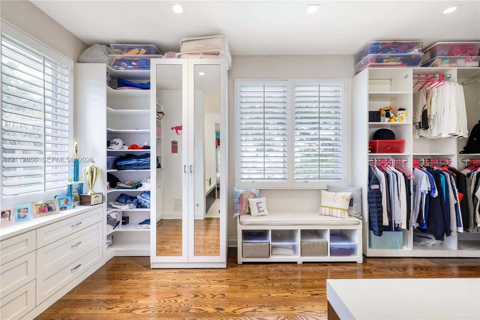 285 Las Brisas Court, Coral Gables, FL 33143 Custom closet featuring white built-in cabinetry, floor-to-ceiling mirrored doors, integrated window seating, plantation shutters, and wood-finish flooring