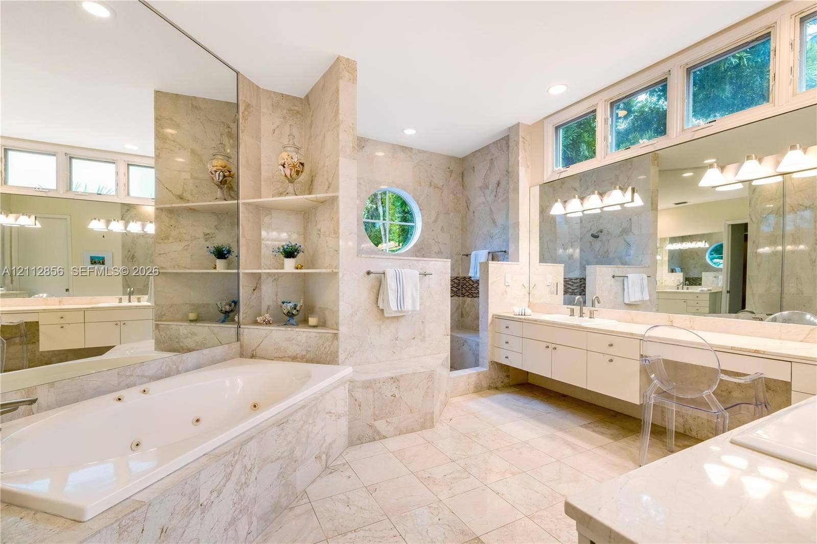 285 Las Brisas Court, Coral Gables, FL 33143 Spacious bathroom featuring stone-tiled surfaces, a built-in jetted tub, a circular accent window, dual vanities with integrated cabinetry, and extensive mirrored wall panels