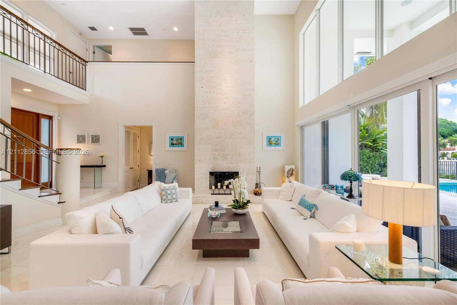 285 Las Brisas Court, Coral Gables, FL 33143 Double-height living area featuring a floor-to-ceiling stone fireplace, expansive glass wall panels, a mezzanine balcony with metal railing, and polished stone flooring