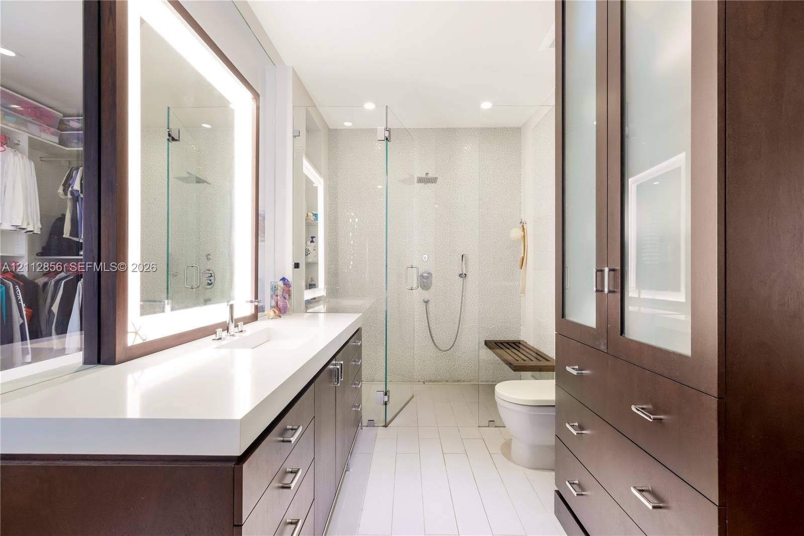285 Las Brisas Court, Coral Gables, FL 33143 Custom dark-wood cabinetry, white composite countertops, integrated LED-backlit mirror, glass-enclosed walk-in shower with pebble-tiled wall, and built-in floor-to-ceiling storage