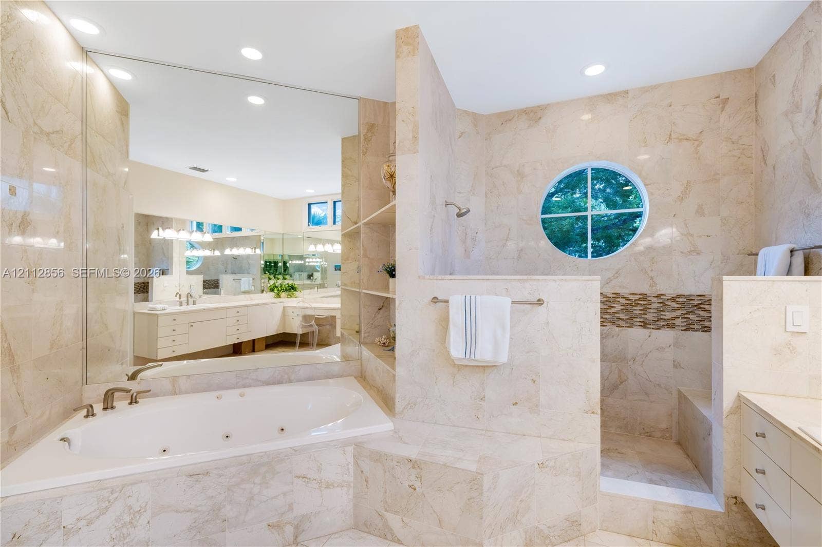 285 Las Brisas Court, Coral Gables, FL 33143 Stone-tiled bathroom featuring a jetted corner tub, walk-in shower with circular window, expansive wall-to-wall mirror, and built-in shelving
