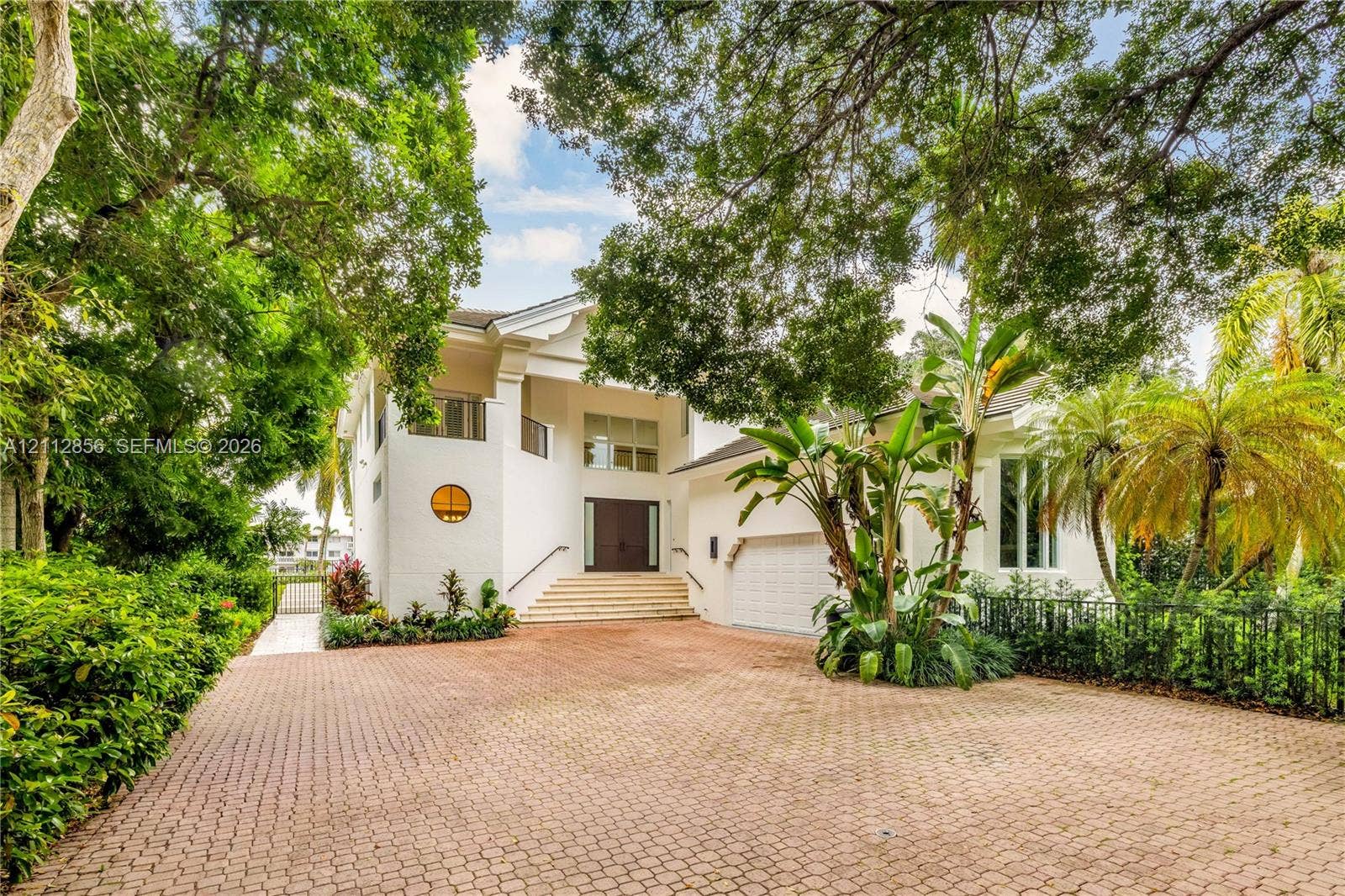 285 Las Brisas Court, Coral Gables, FL 33143 Stucco exterior featuring a circular accent window, elevated entryway with stone steps, multi-car garage, and a spacious paved driveway shaded by mature trees
