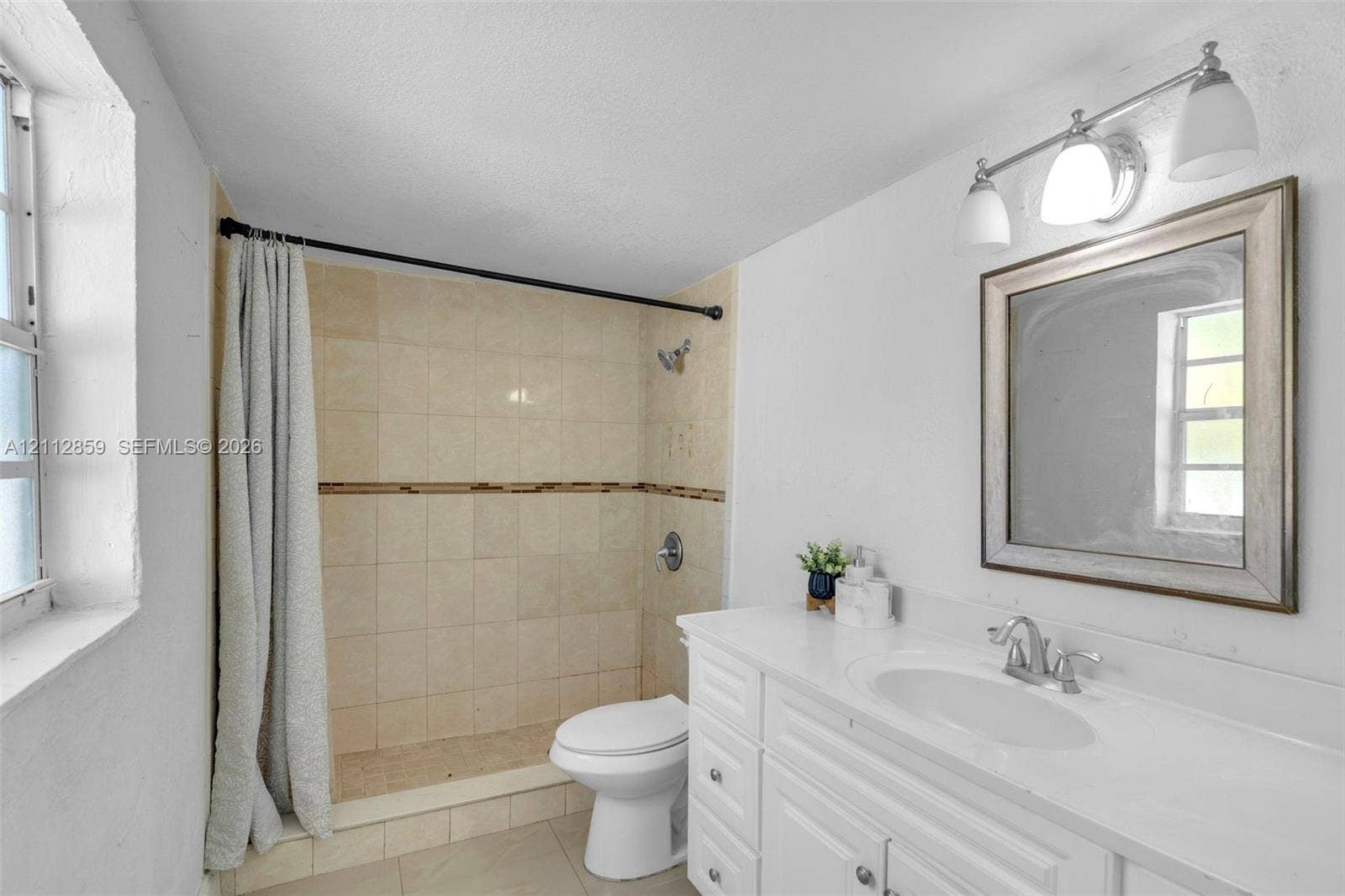 1207 Chateau Park Drive, Fort Lauderdale, FL 33311 Bathroom featuring a white vanity with an integrated sink, a rectangular framed mirror, a triple-light fixture, and a tiled shower stall with a decorative accent border
