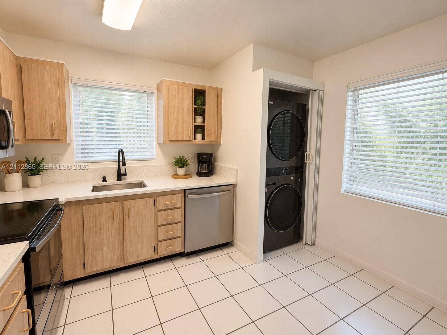 Kitchen featuring light wood-finish cabinetry, white solid-surface countertops, stainless steel undermount sink with matte black faucet, ceramic tile flooring, and a built-in laundry closet