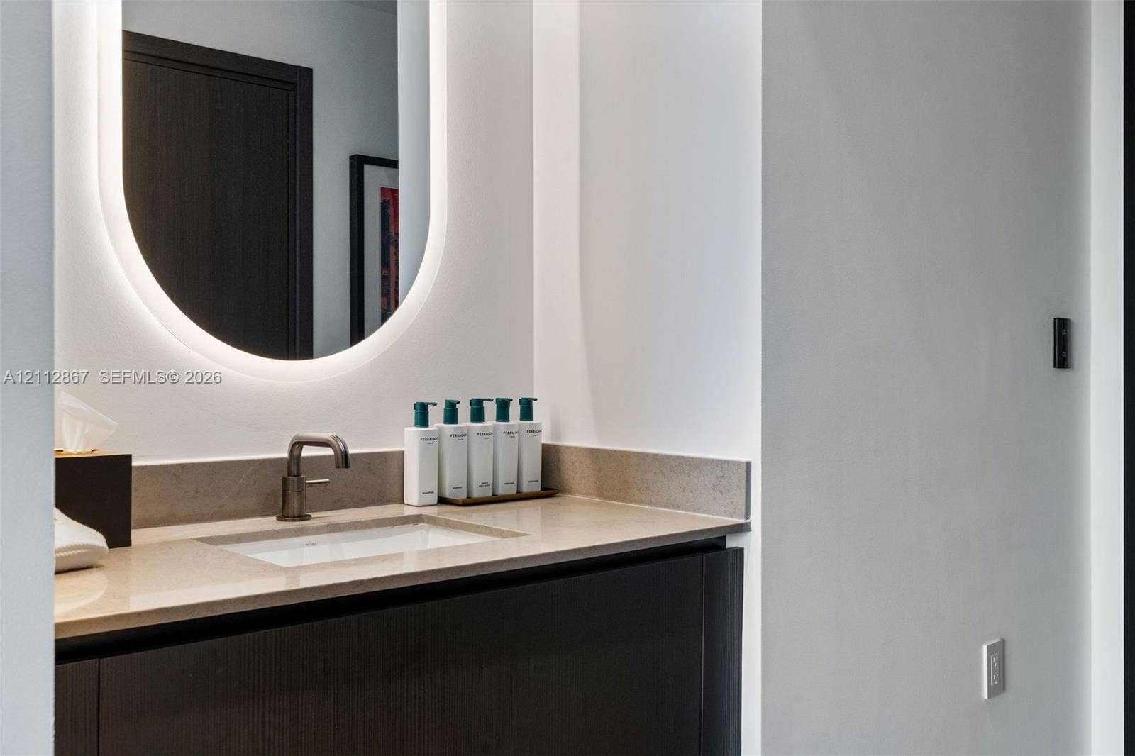 Dark-finish cabinetry with a neutral stone countertop, integrated rectangular sink, wall-mounted metal faucet, and backlit oval mirror