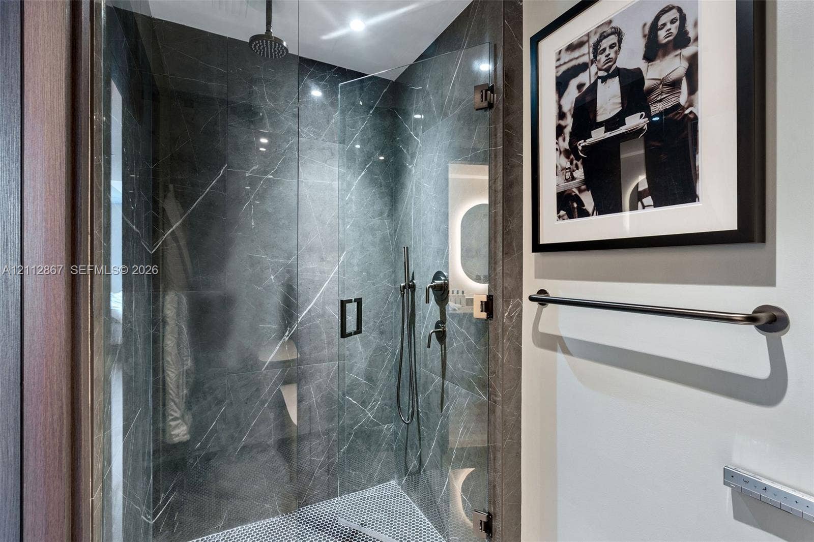 Walk-in shower enclosure featuring full-height stone tile surround, frameless glass door, rain-style showerhead, hand-held sprayer, and dark-finish metal hardware