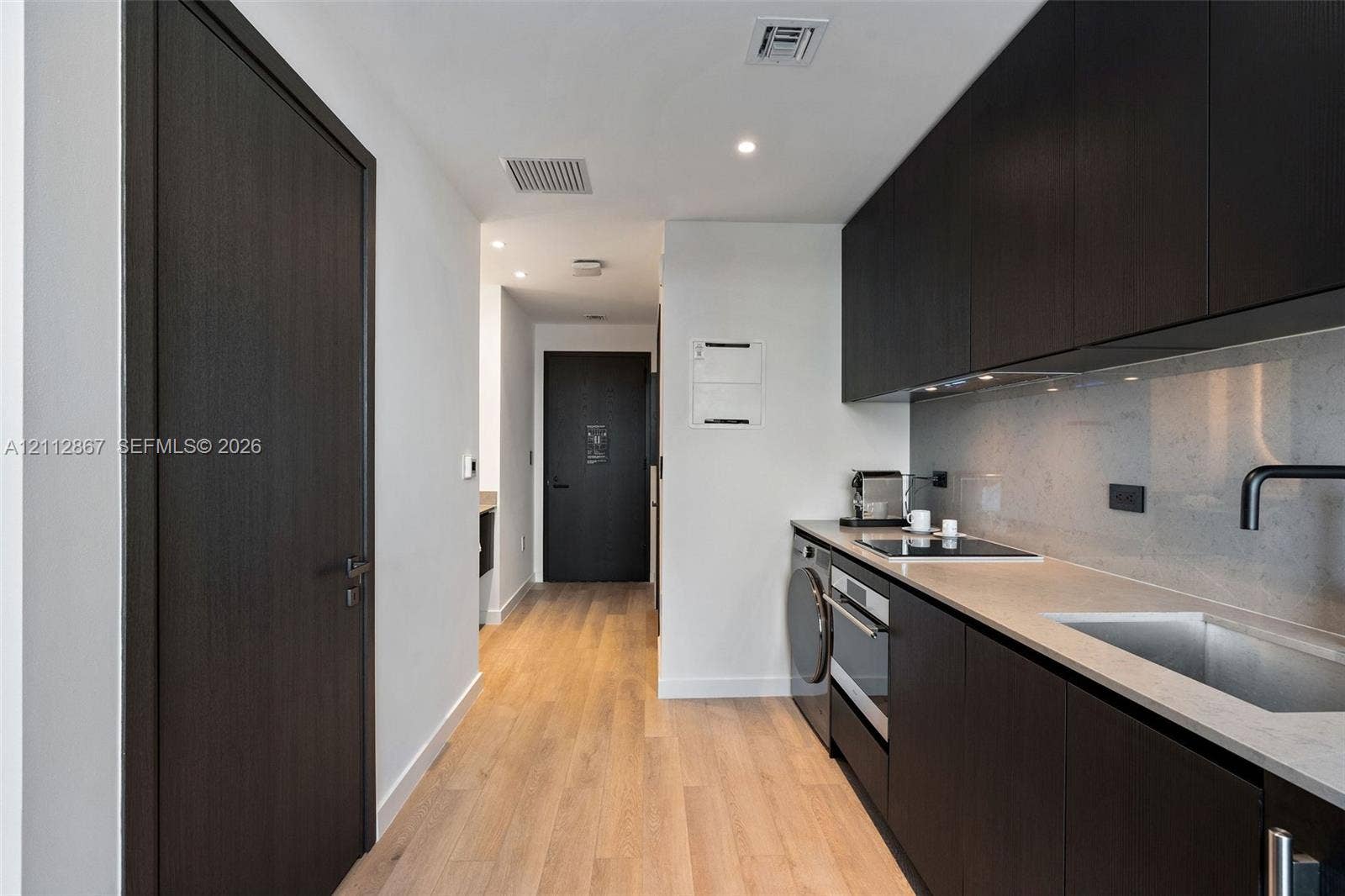 Kitchenette featuring dark-finish cabinetry, solid-surface countertops, integrated cooktop and oven, under-mount sink with matte black faucet, and wood-finish flooring