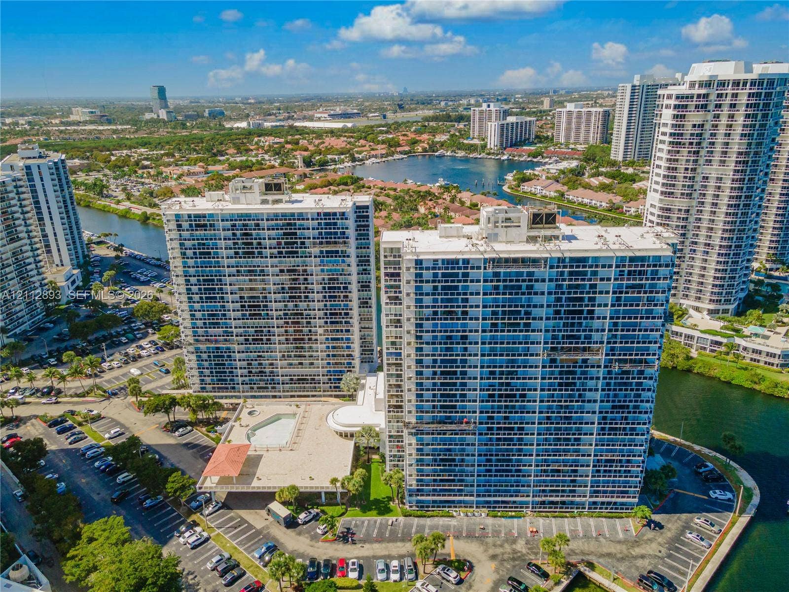 20515 E Country Club Drive, #1048, Aventura, FL 33180 High-rise residential towers featuring expansive glass facades, a ground-level swimming pool, surface parking, and waterfront positioning