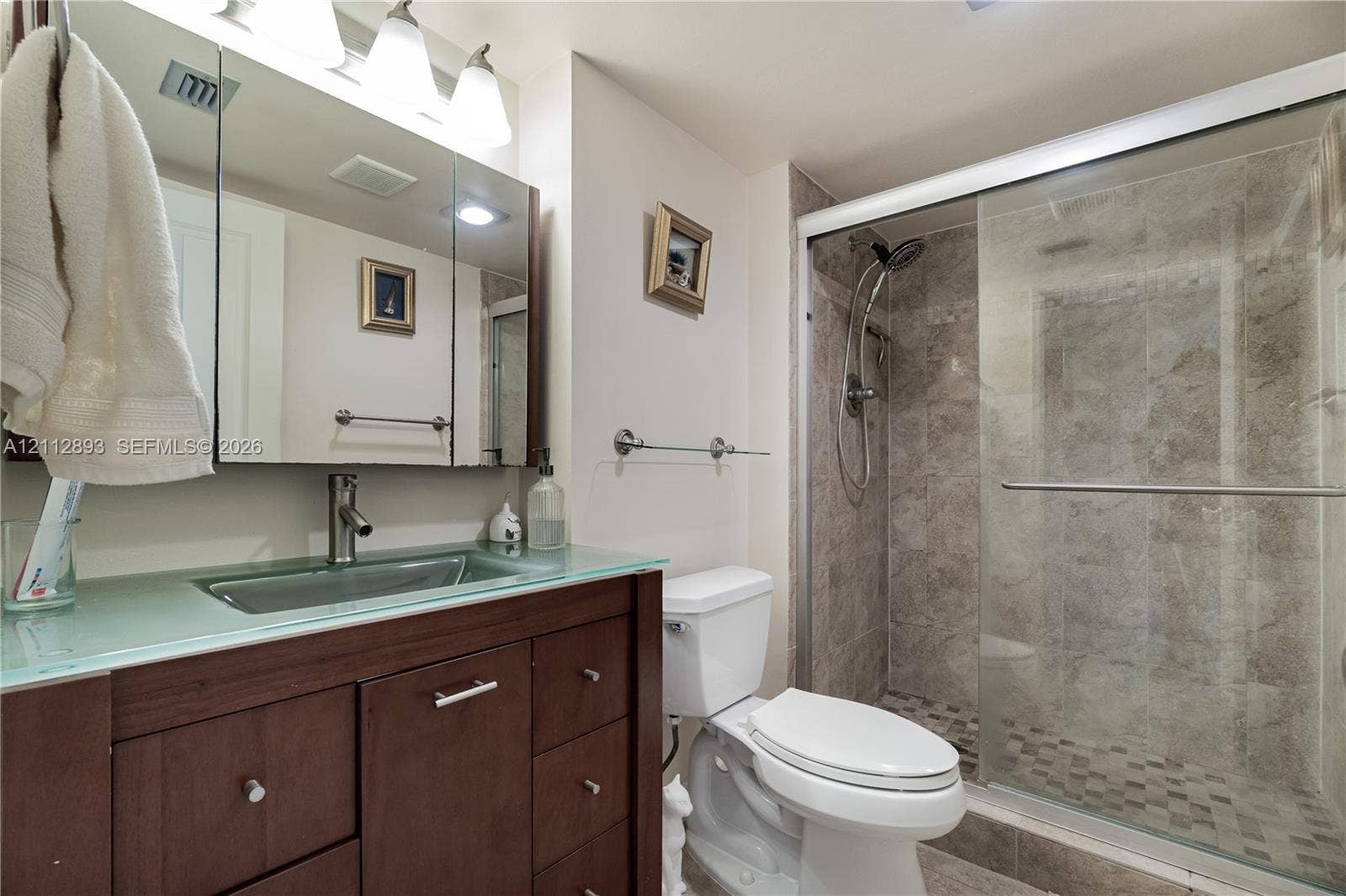 20515 E Country Club Drive, #1048, Aventura, FL 33180 Vanity featuring a glass countertop with an integrated rectangular sink, dark wood-finish cabinetry, mirrored medicine cabinet, and a walk-in shower with tiled walls and glass sliding doors