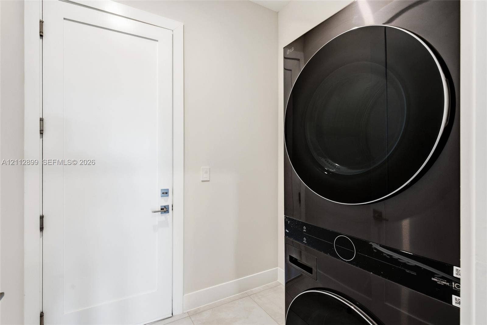 2747 SW 23rd Terrace, Miami, FL 33145 Stacked washer and dryer unit with a sleek black finish, paired with a white Shaker-style door featuring polished chrome hardware and large-format tile flooring
