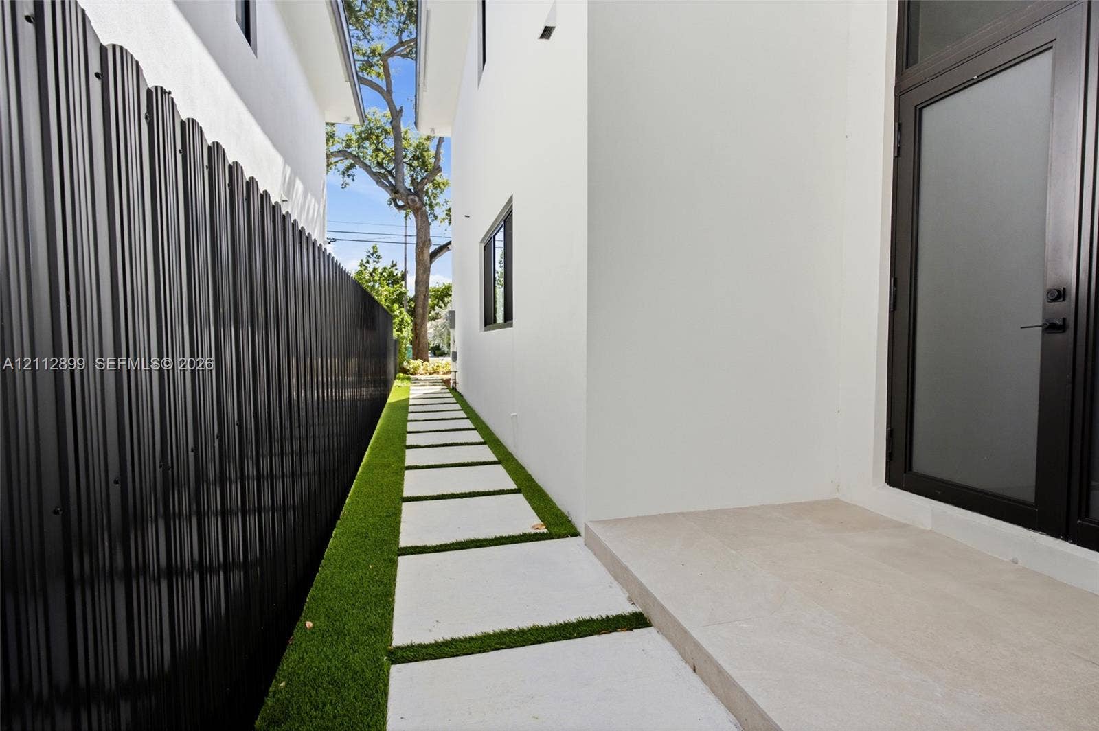 2747 SW 23rd Terrace, Miami, FL 33145 Side exterior featuring white stucco siding, a concrete paver walkway set in artificial turf, a black metal corrugated privacy fence, and a glass-panel entry door