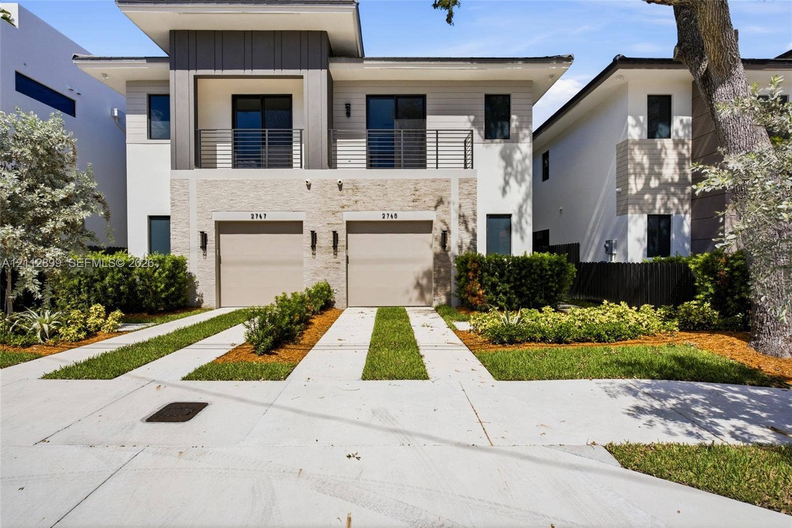 2747 SW 23rd Terrace, Miami, FL 33145 Modern exterior featuring a stacked stone facade, metal-accented second-story balcony with railings, dual two-car garage doors, and concrete paver driveways with integrated grass strips