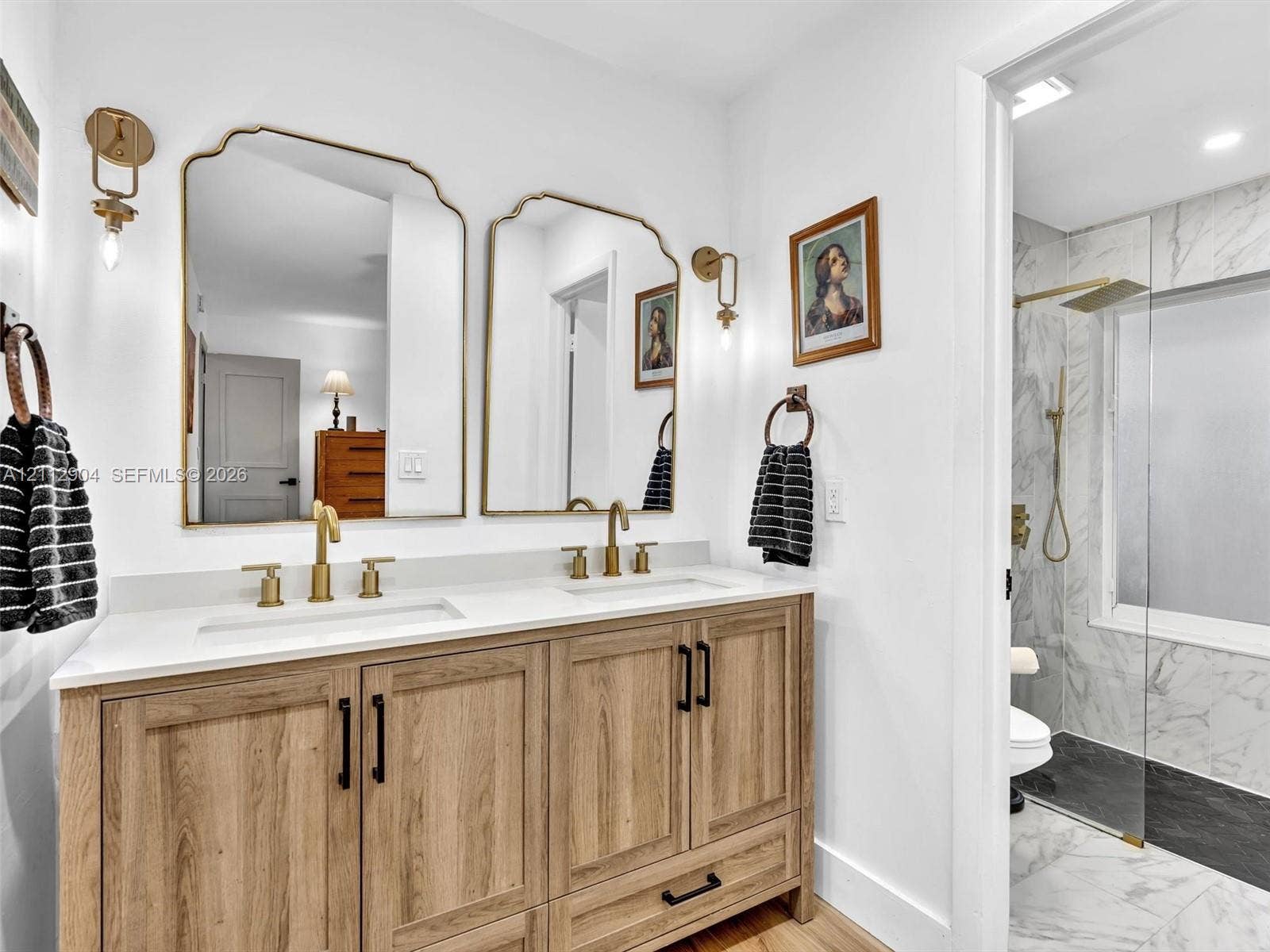 76 Forest Circle, Cooper City, FL 33026 Dual-sink vanity with wood-finish cabinetry, quartz countertop, brass fixtures, arched metal-frame mirrors, and a glass-enclosed shower