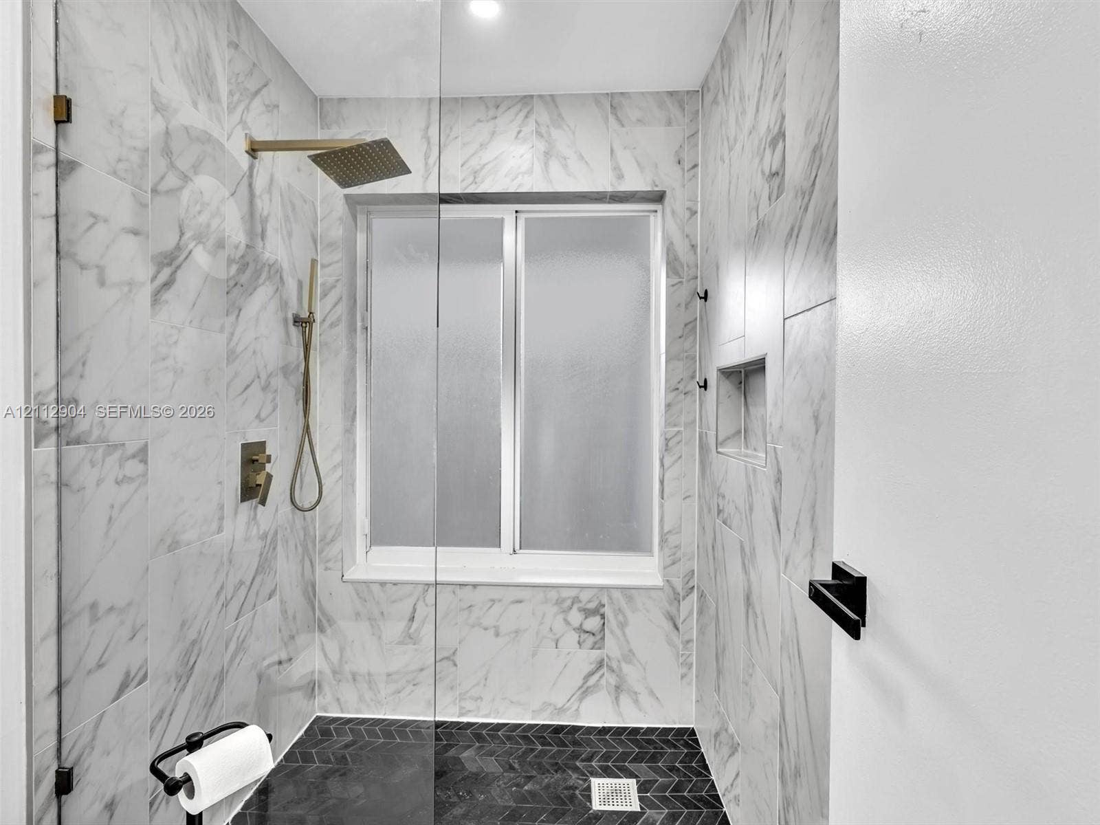 76 Forest Circle, Cooper City, FL 33026 Walk-in shower featuring large-format stone-finish wall tile, herringbone-patterned floor tile, a recessed wall niche, brass-finish rainfall shower head with handheld attachment, and a glass partition